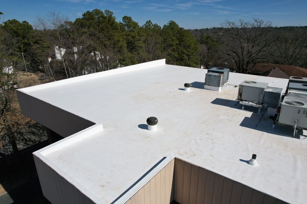 A flat white roof on a building with HVAC units and vents, surrounded by trees under a clear blue sky.