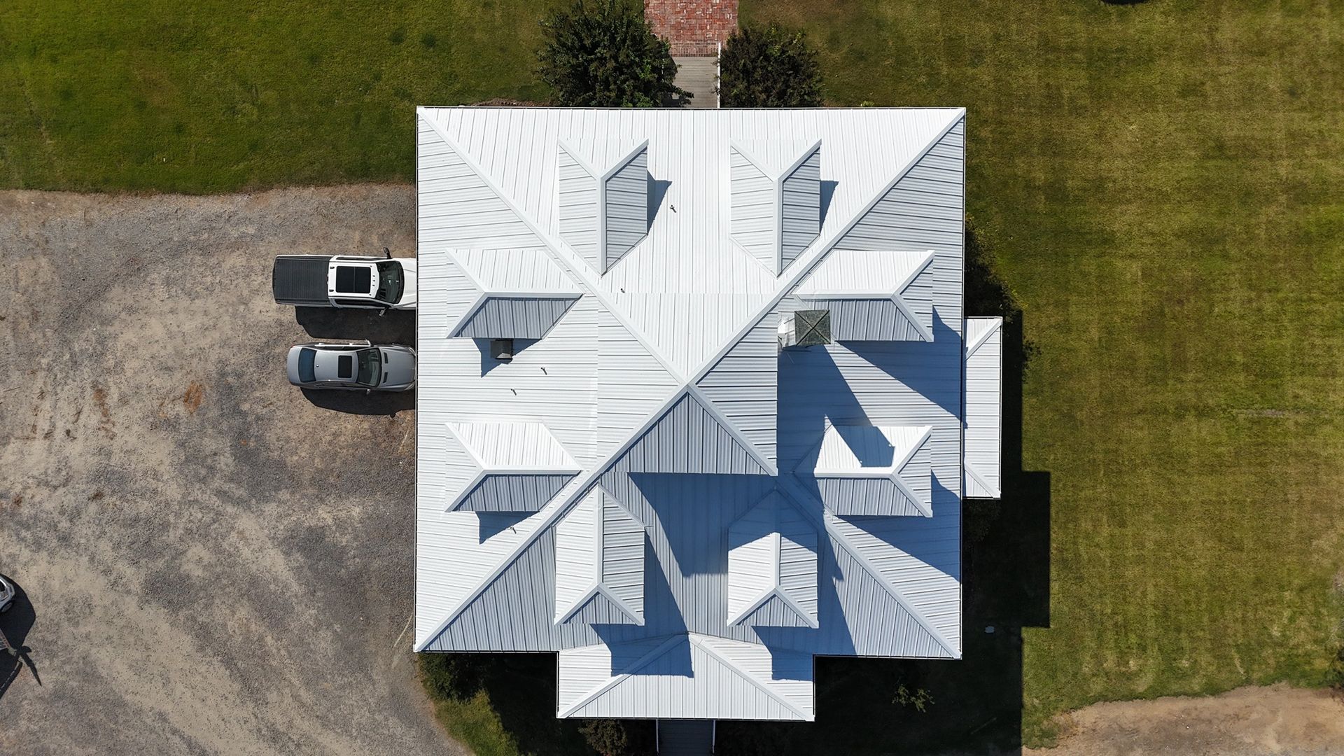 An aerial top-down view of a house with a white metal roof, two cars parked on a gravel driveway, and surrounding lawn.