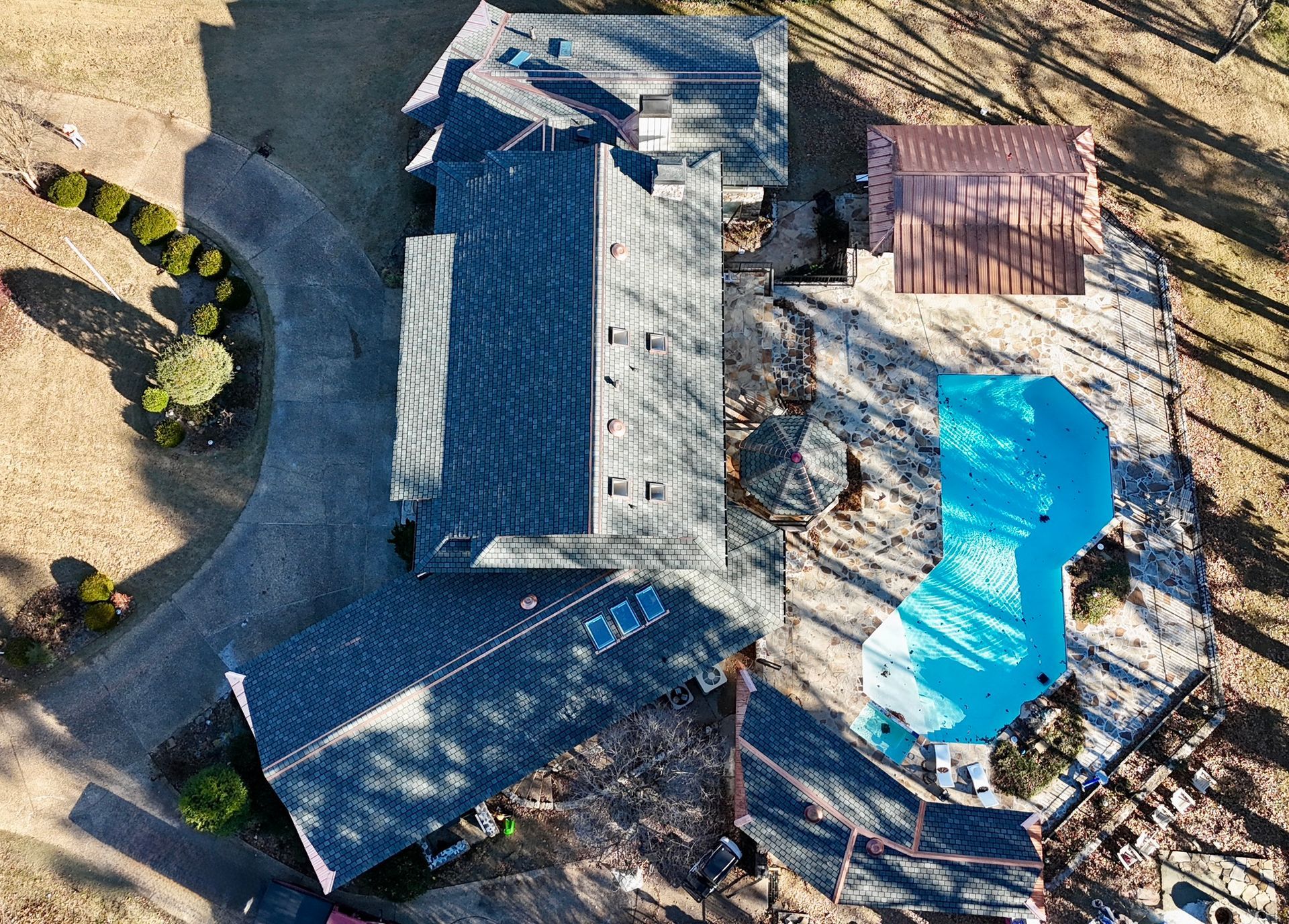 Aerial view of a house with a dark shingled roof, a blue swimming pool, and a pergola on a stone patio in a wooded yard.