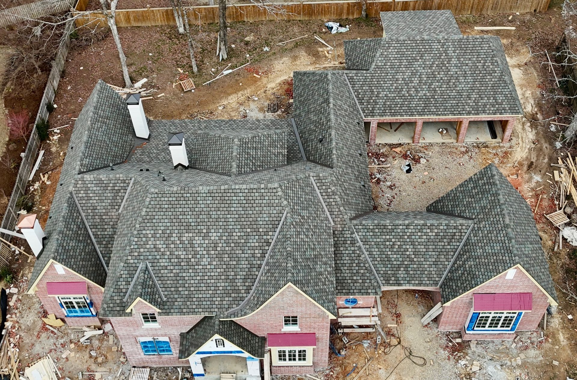 Aerial view of a partially constructed brick house with a complex, shingled grey roof, set on a dirt residential lot.