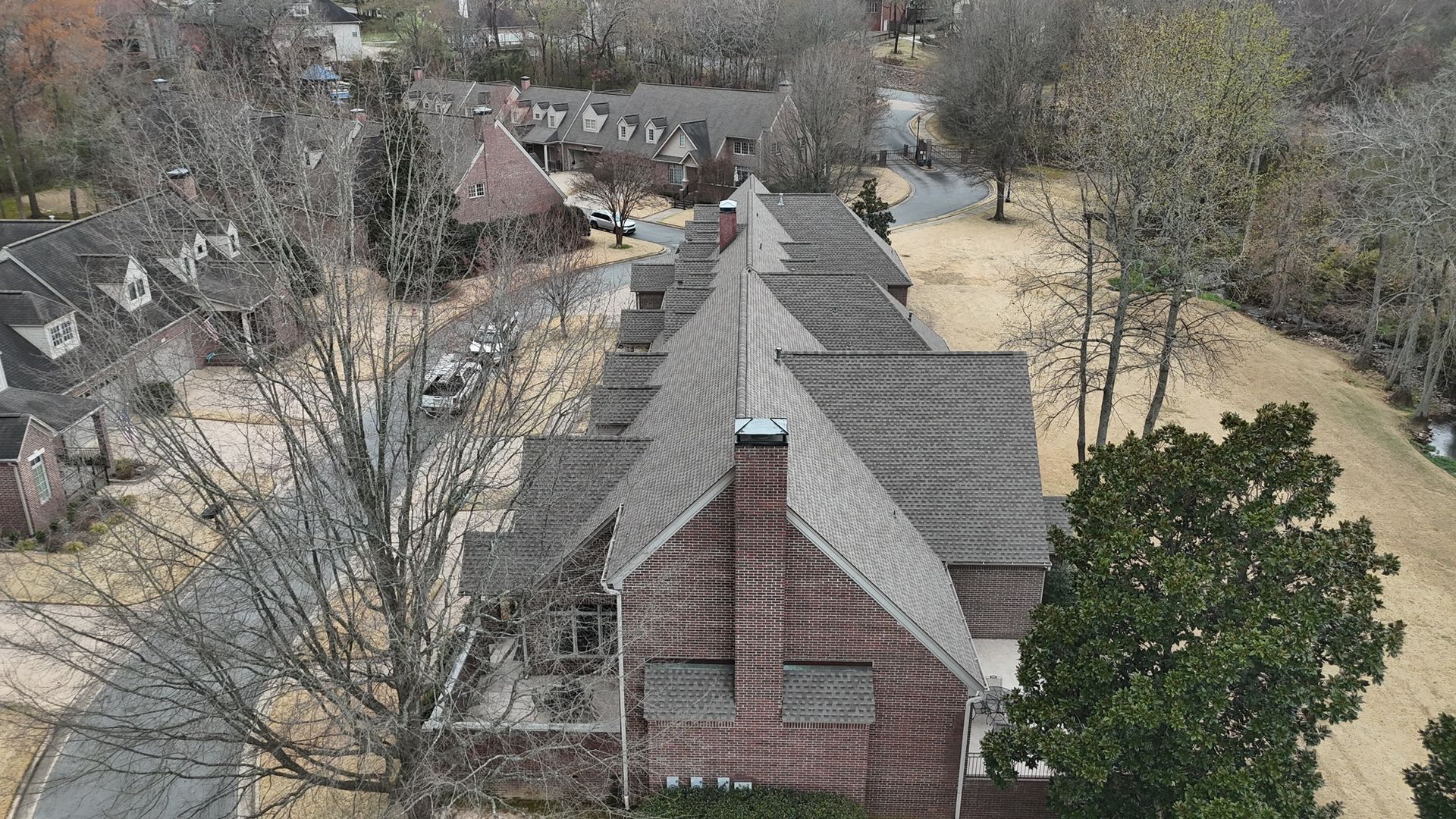 Aerial view of a brown brick house with a gray shingled roof, surrounded by trees and a curving driveway.