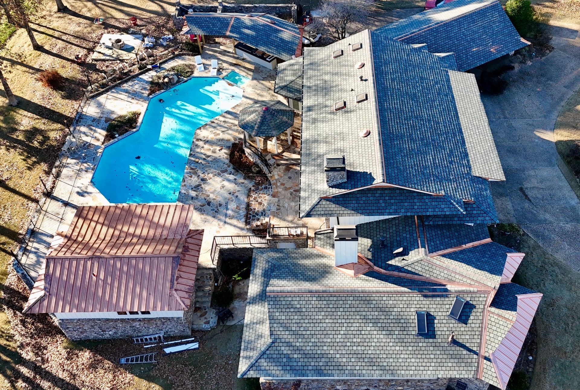 Aerial view of a home with a large blue swimming pool, dark shingled roof, and a separate copper-roofed structure.