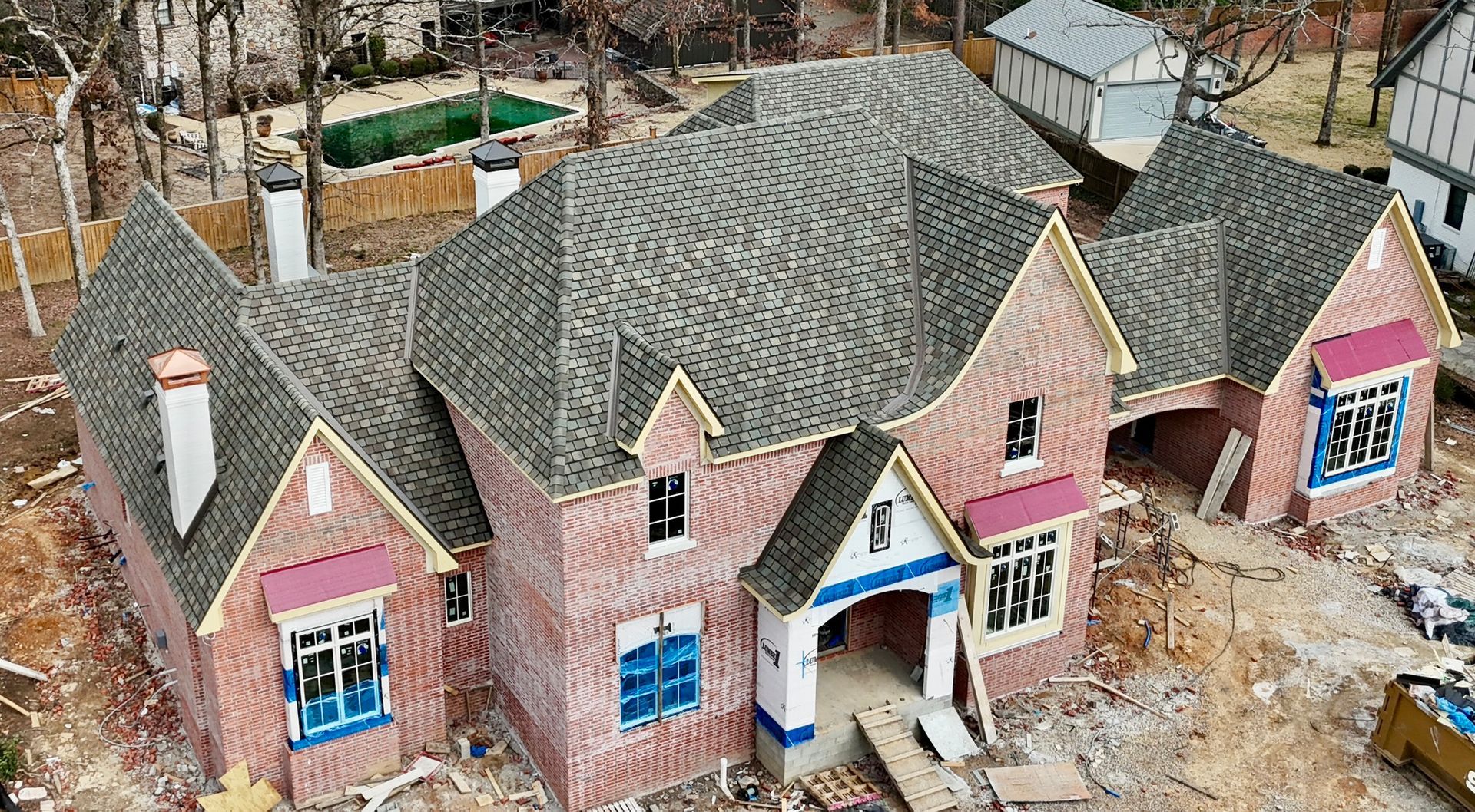 A high-angle view of a brick house under construction, featuring gray shingled roofs and windows covered in blue film.