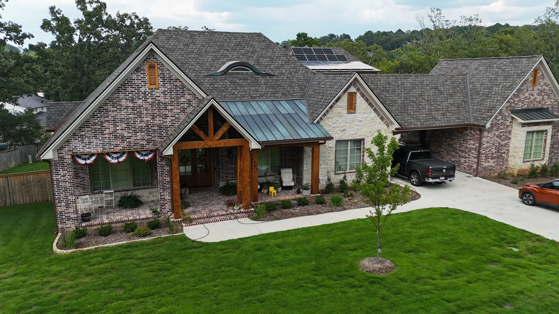 A stone suburban house with a dark metal roof, wood entryway, green lawn, and a driveway with parked vehicles.