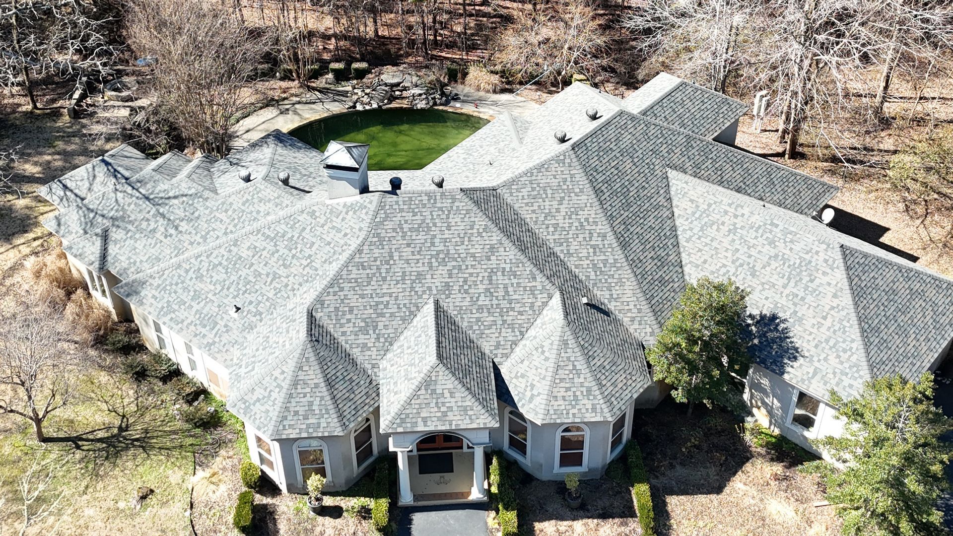 An aerial view of a sprawling suburban home with a multi-gabled gray shingle roof, set among trees and a circular lawn.