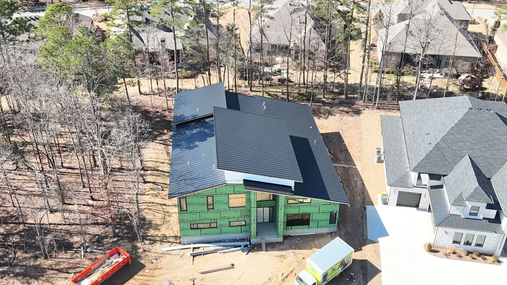 An aerial view of a construction site featuring a green-sheathed house frame with a finished black metal roof.