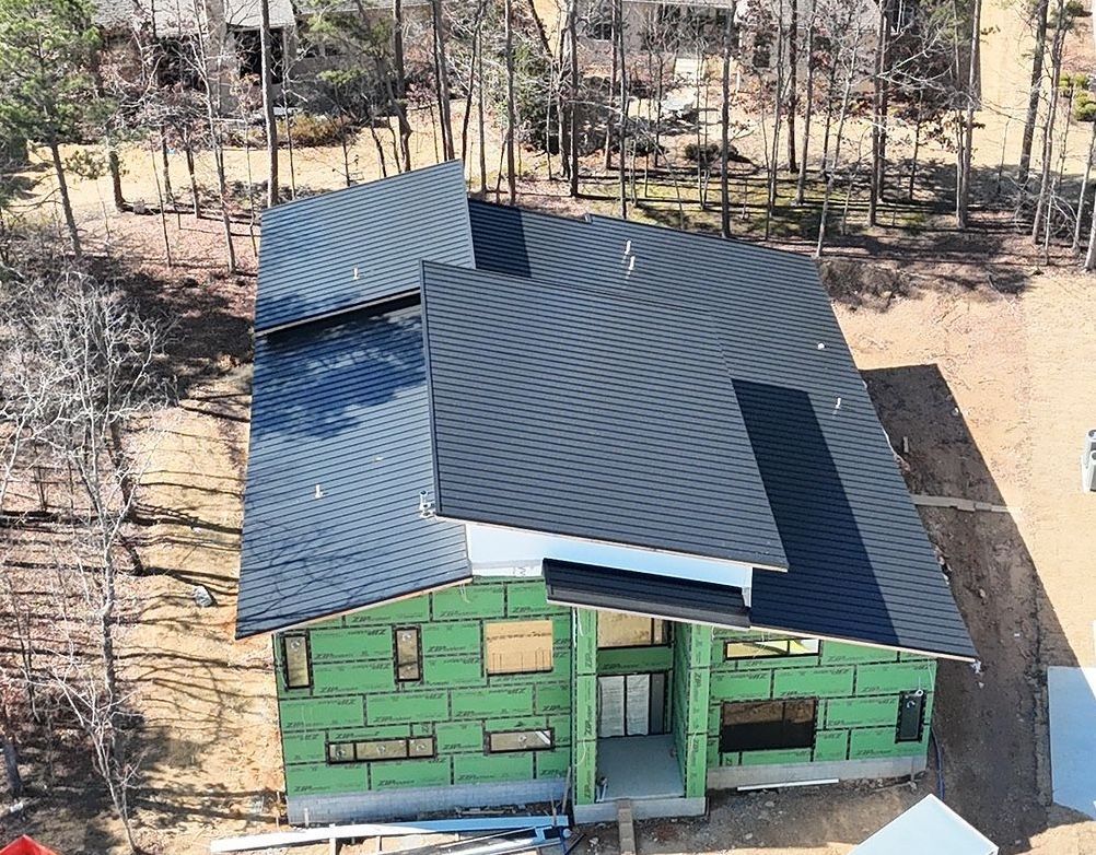 An aerial view of a house under construction, featuring a dark metal roof, green sheathing, and a wooded exterior.