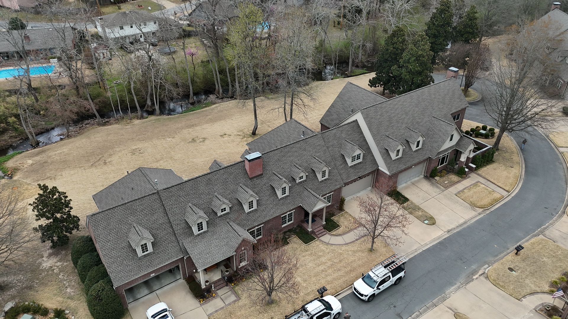 Aerial view of a large multi-roofed brick home with a winding driveway and two service vehicles parked in front.