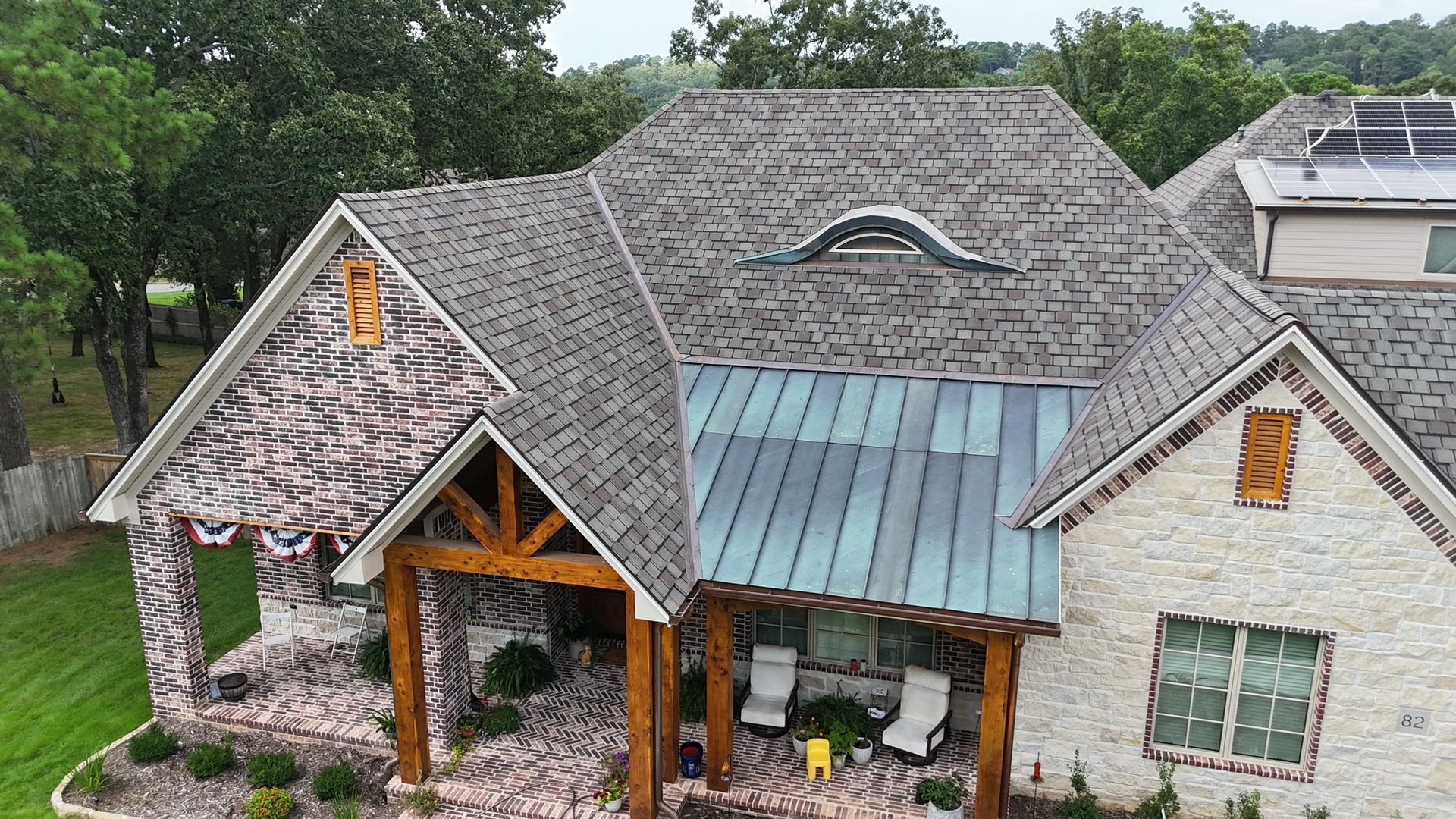 High-angle view of a brick house with a multi-gabled shingle roof, a copper-paneled awning, and a wooden porch.