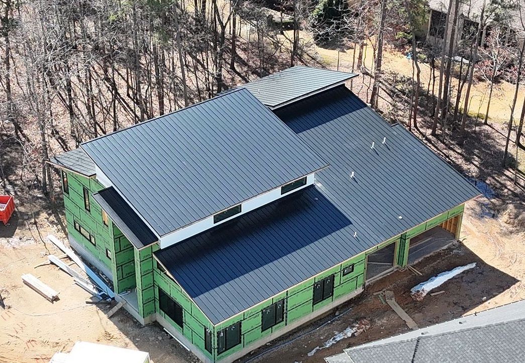 A high-angle view of a house under construction with a black standing-seam metal roof and green exterior sheathing.