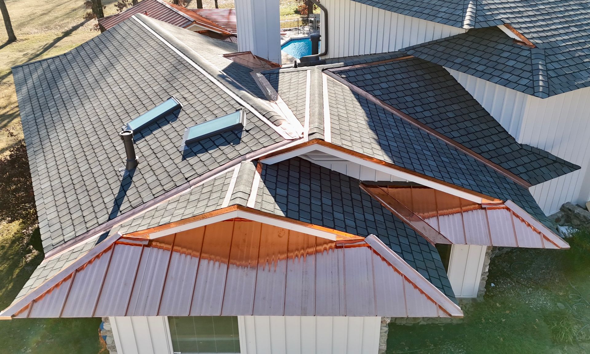 An aerial view of a house roof featuring dark grey shingles, copper metal accents, and two rectangular skylights.