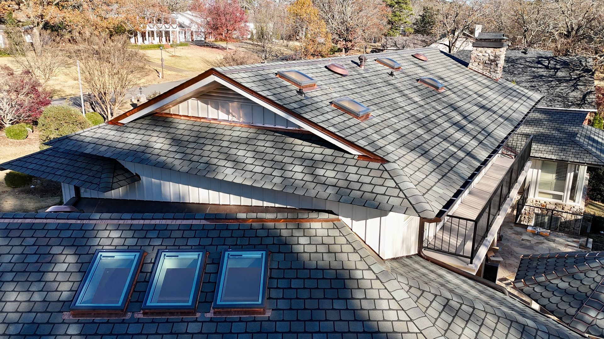 Aerial view of a gray shingled roof with several skylights, copper flashing, and trees in the background.