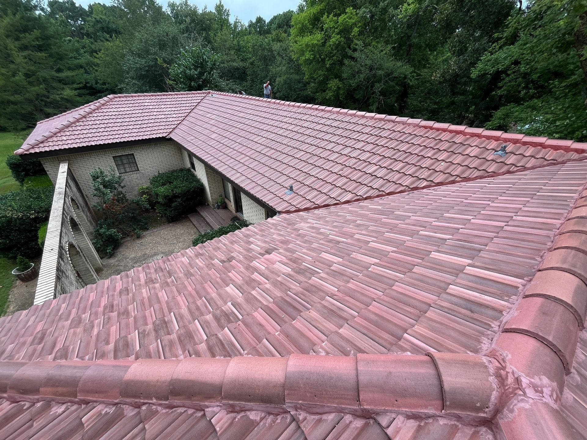 An angled view of a house roof with reddish-brown terracotta tiles, seen from above with trees in the background.