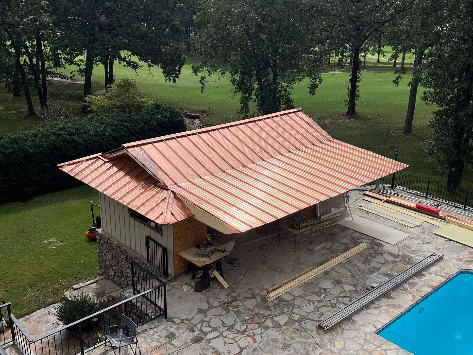 An aerial view shows a pool house with a newly installed copper-colored metal roof, situated next to a swimming pool.