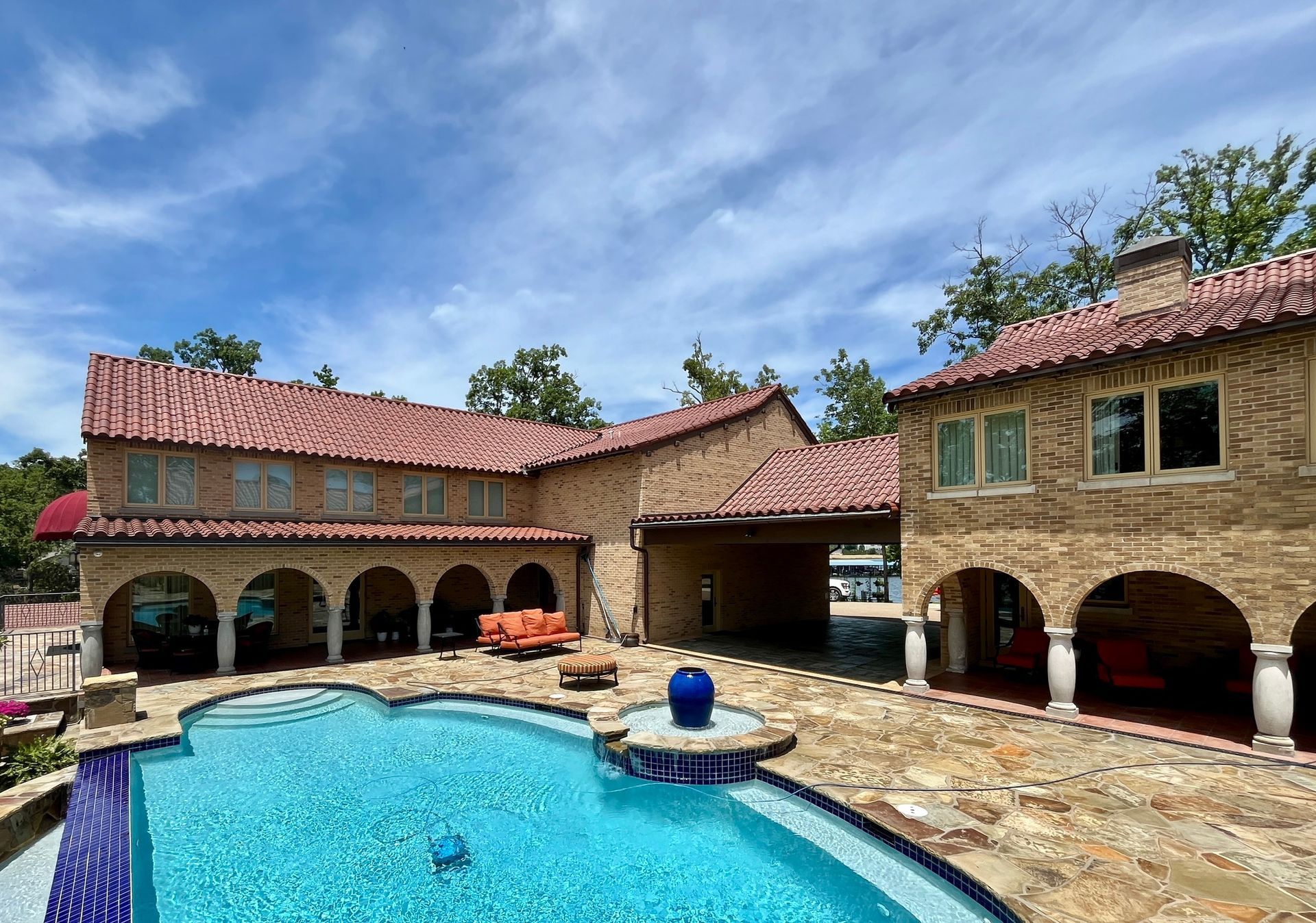 A multi-story stone house with a red tile roof and arched patio, overlooking a blue swimming pool under a sunny sky.