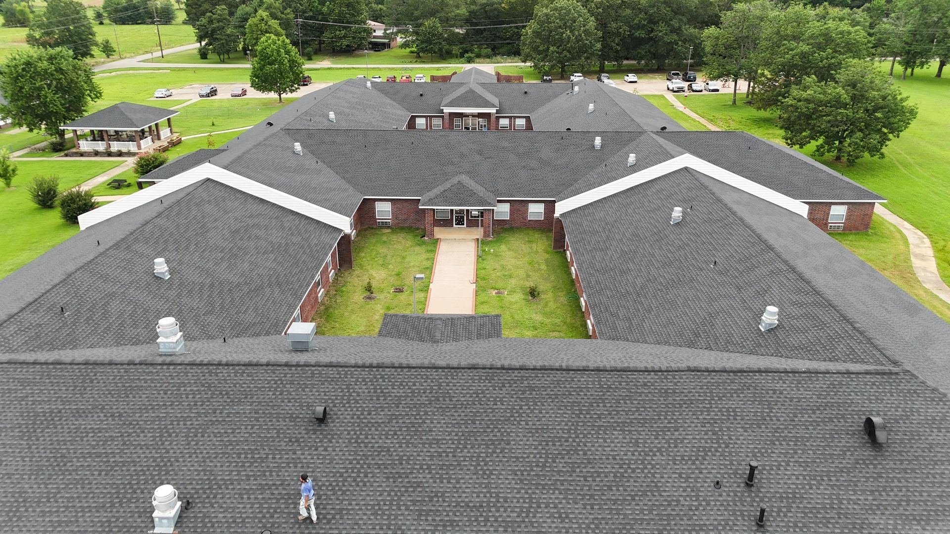 Aerial view of a U-shaped brick building with a dark shingled roof, surrounded by a green lawn and trees.