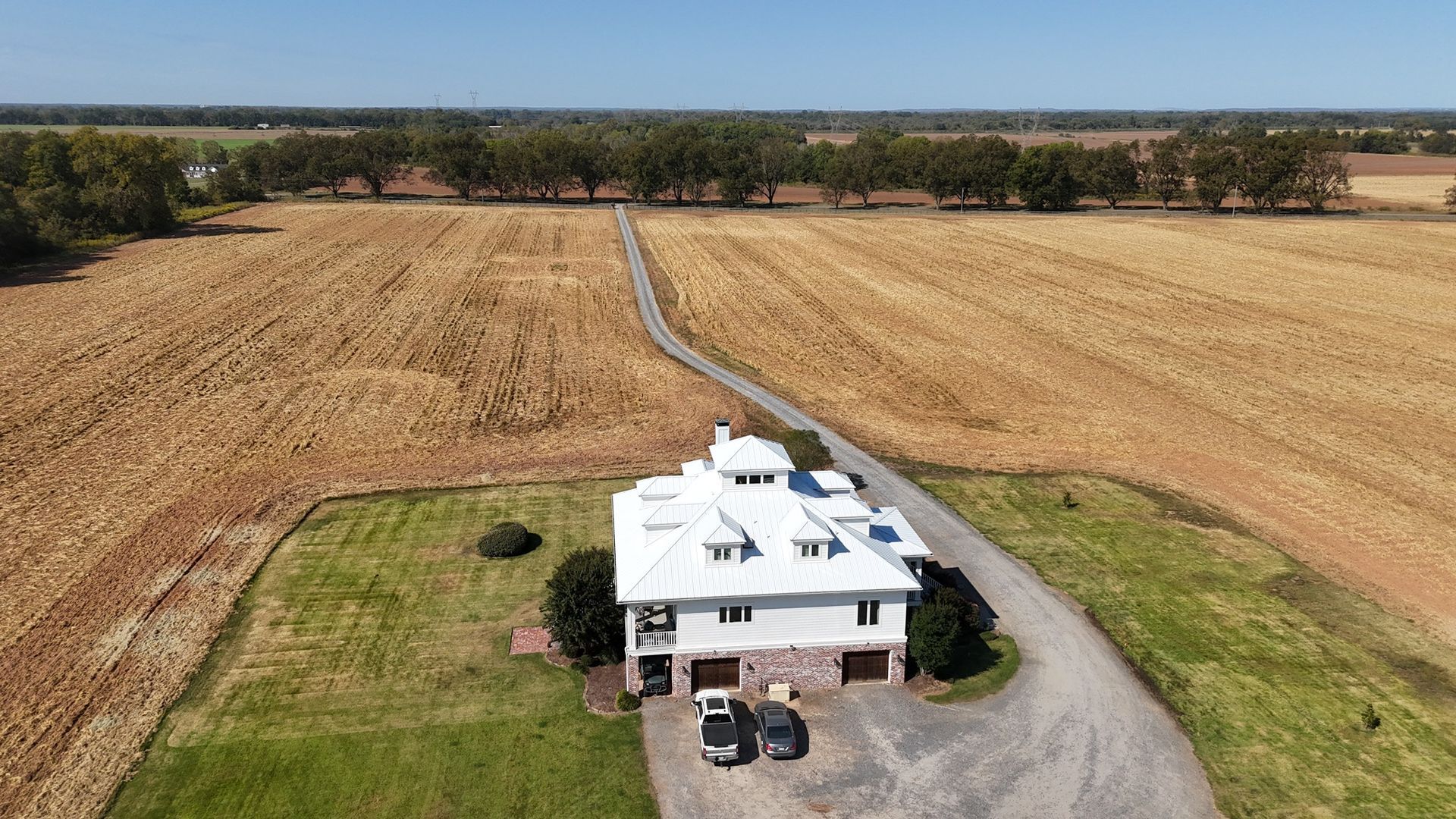 An aerial view of a white two-story house with a metal roof, situated in the middle of a large, harvested agricultural field.