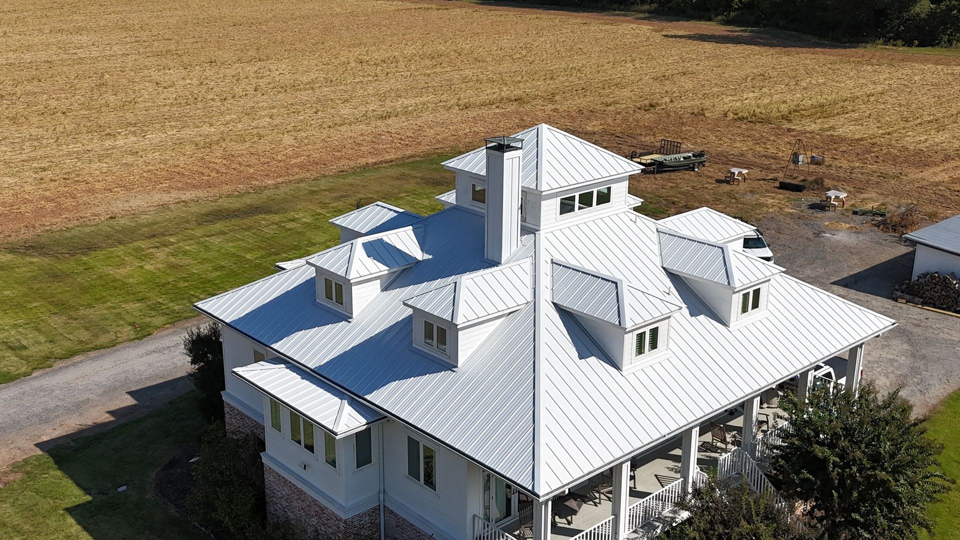 An aerial view of a white farmhouse with a metal roof, dormer windows, and a tall central chimney set in a rural landscape.