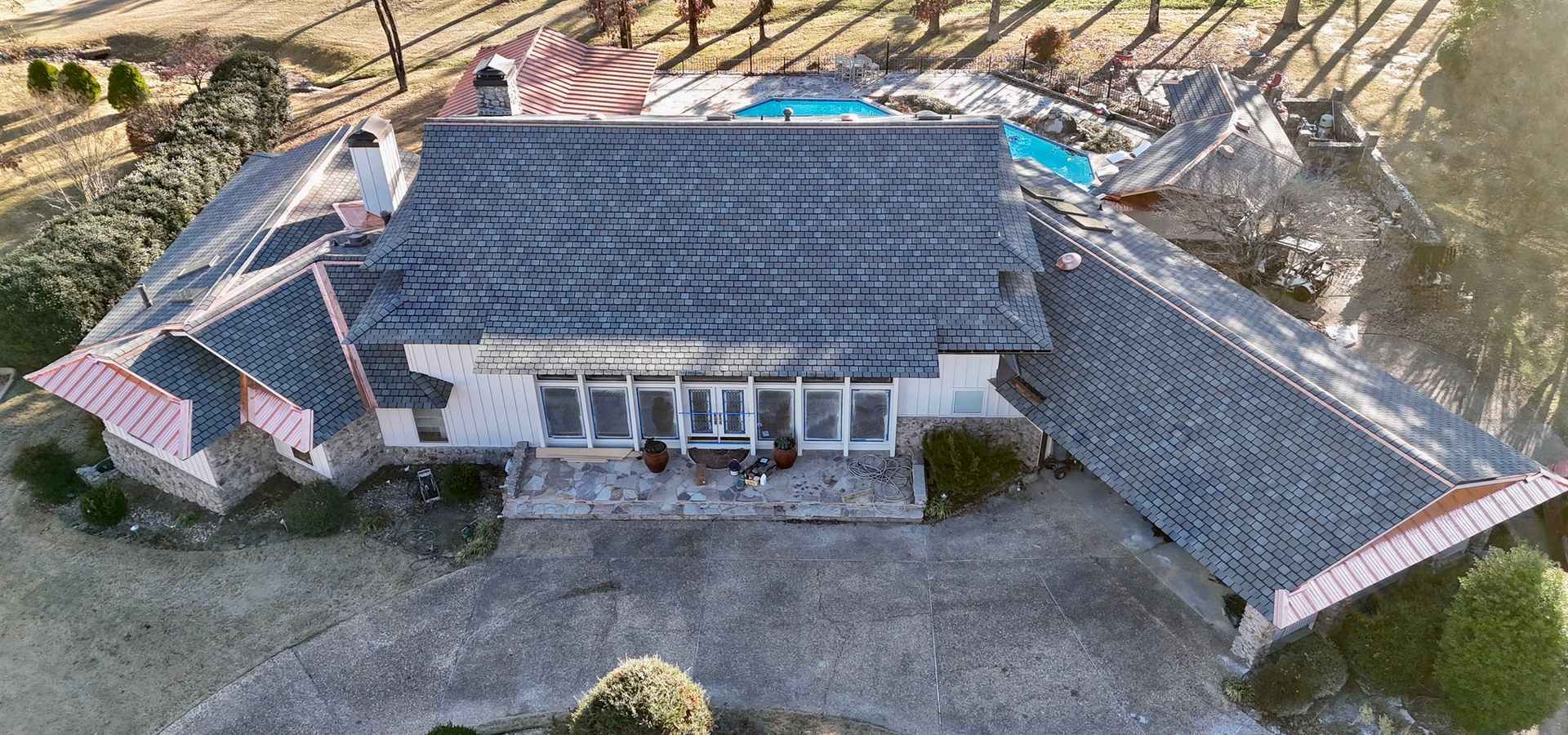 An aerial view of a white single-story house with a dark shingled roof, surrounded by trees and a pool in a yard.