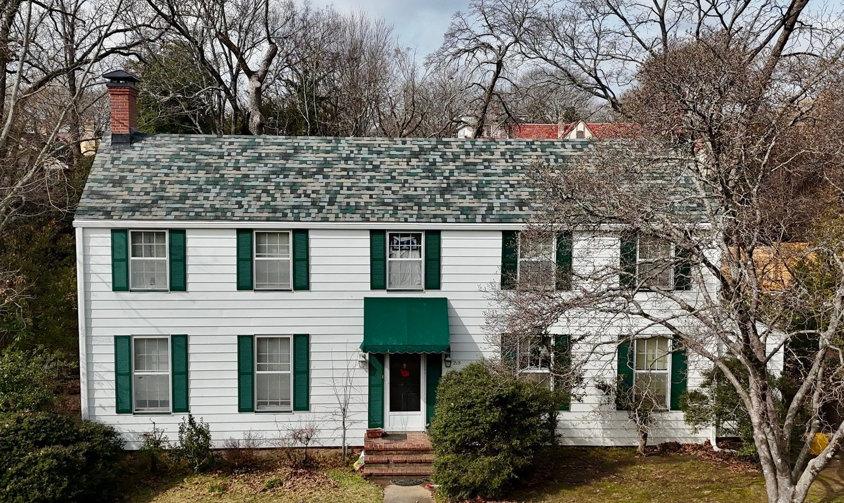 A two-story white house with dark green shutters, a green awning over the front door, and a grey roof among bare trees.