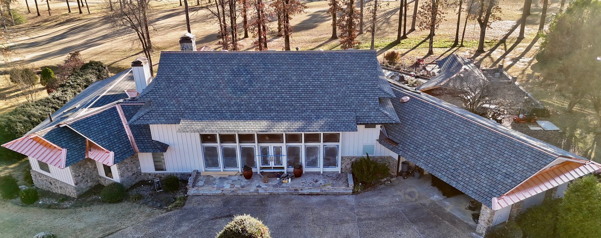 Aerial view of a white, single-story house with a grey shingled roof, stone accents, and a large front patio.