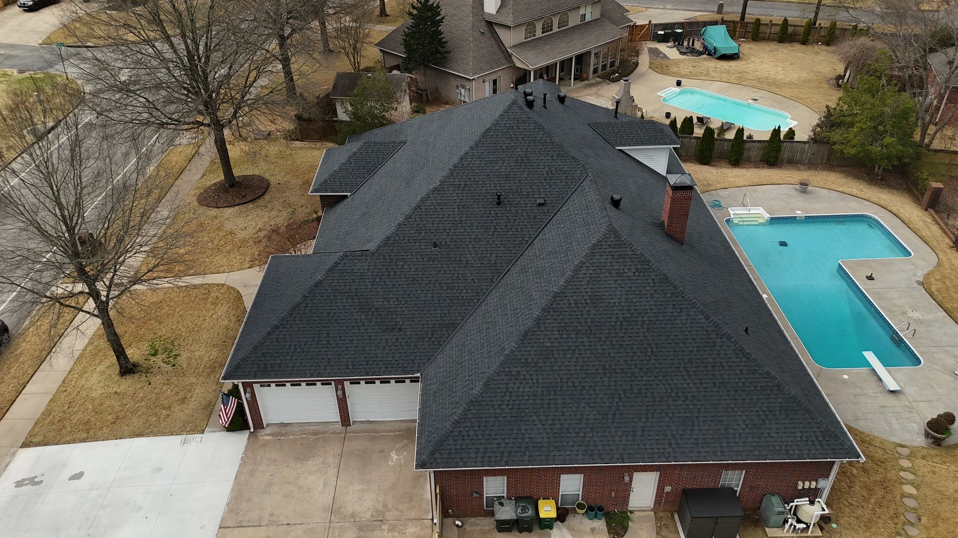 An aerial view of a single-story brick house with a dark shingled roof, attached garage, and a backyard swimming pool.