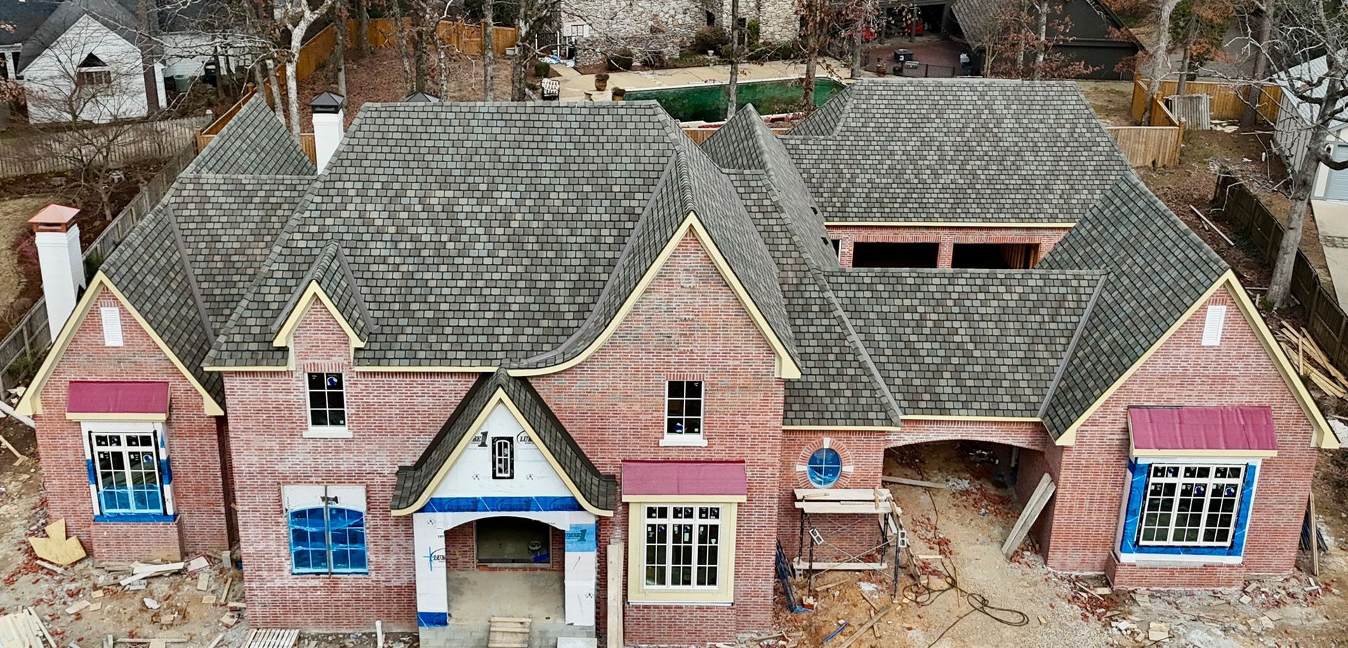 A large, multi-gabled, under-construction brick house with a dark shingled roof seen from an aerial perspective.