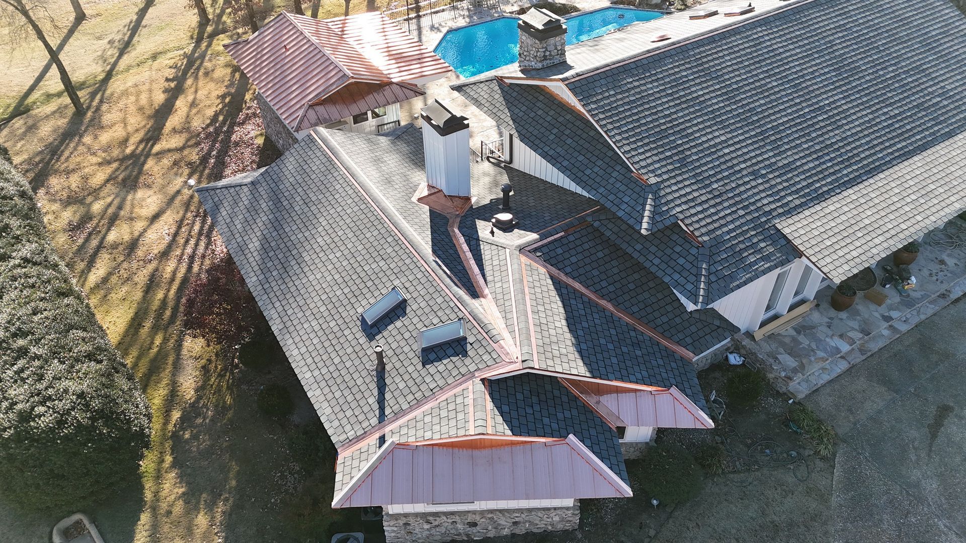 An aerial view of a house roof featuring gray shingles and prominent copper flashing, with a swimming pool in the yard.