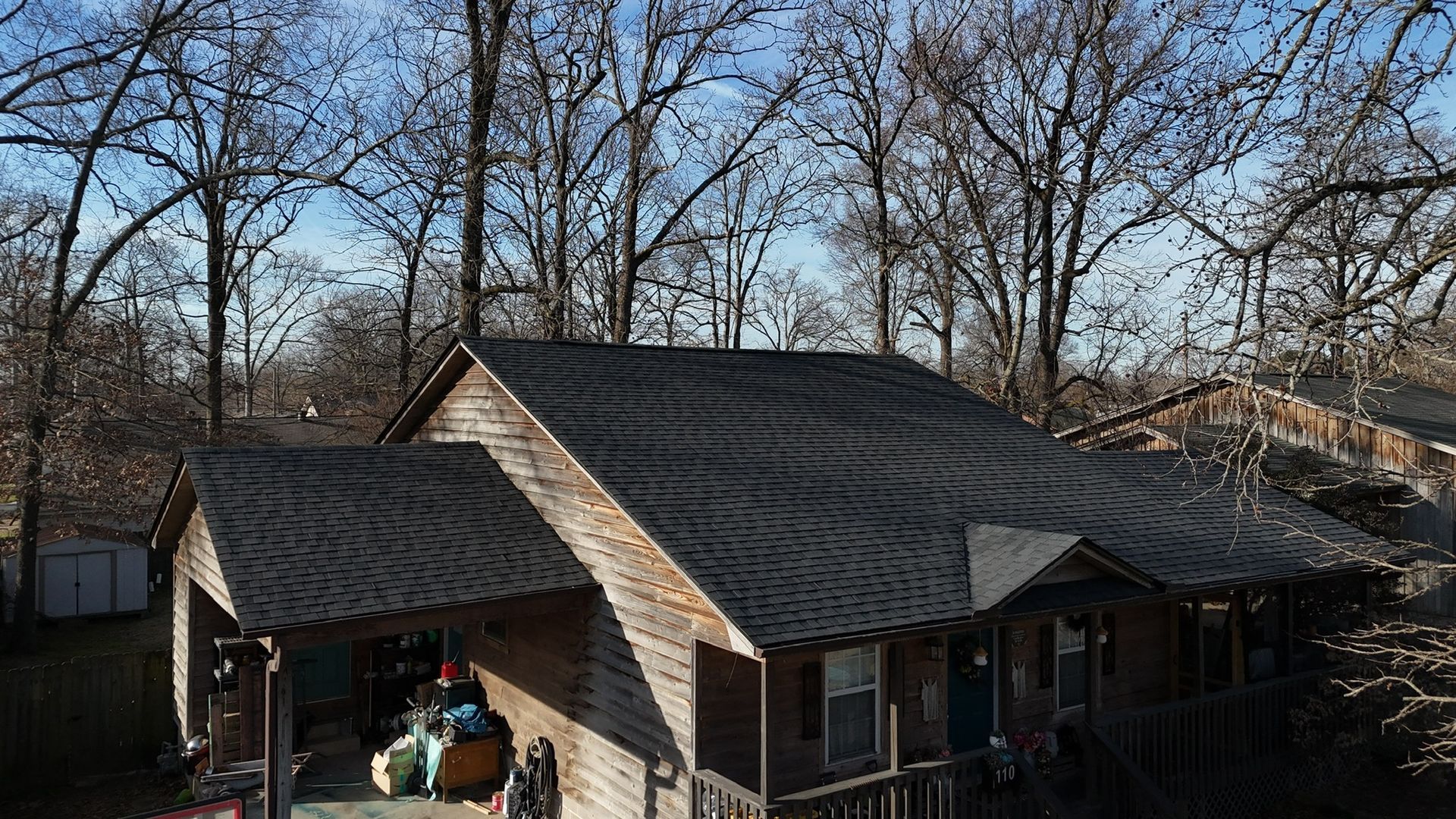 An elevated view of a house with a newly installed dark shingle roof, set among bare trees on a clear, sunny day.