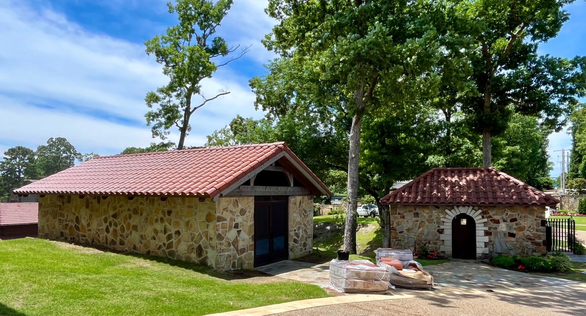 Two rustic stone buildings with red tile roofs sit in a sunny, tree-lined yard under a blue sky.