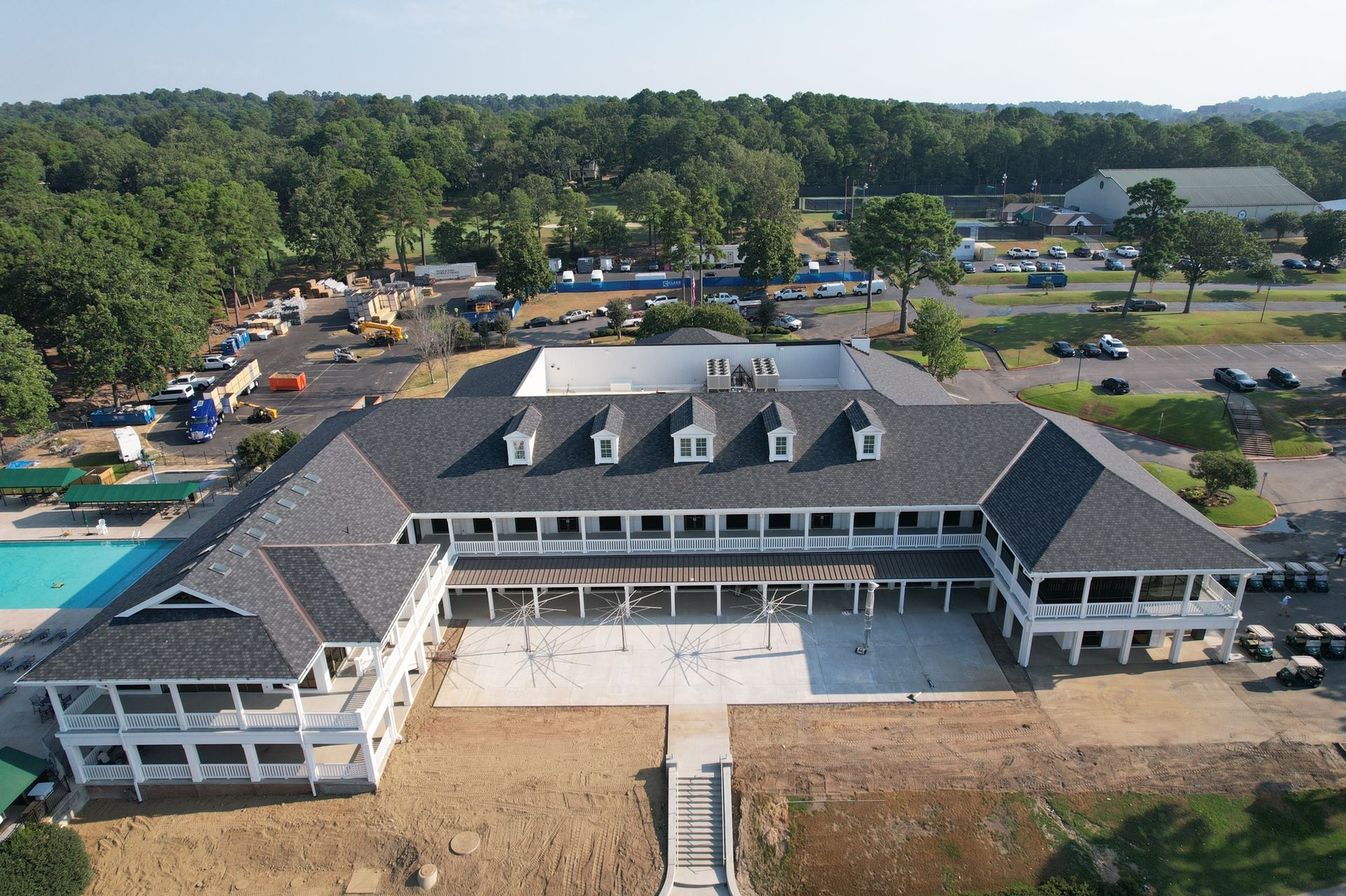 Aerial view of a white, U-shaped building with a gray roof and surrounding paved grounds near a swimming pool and trees.