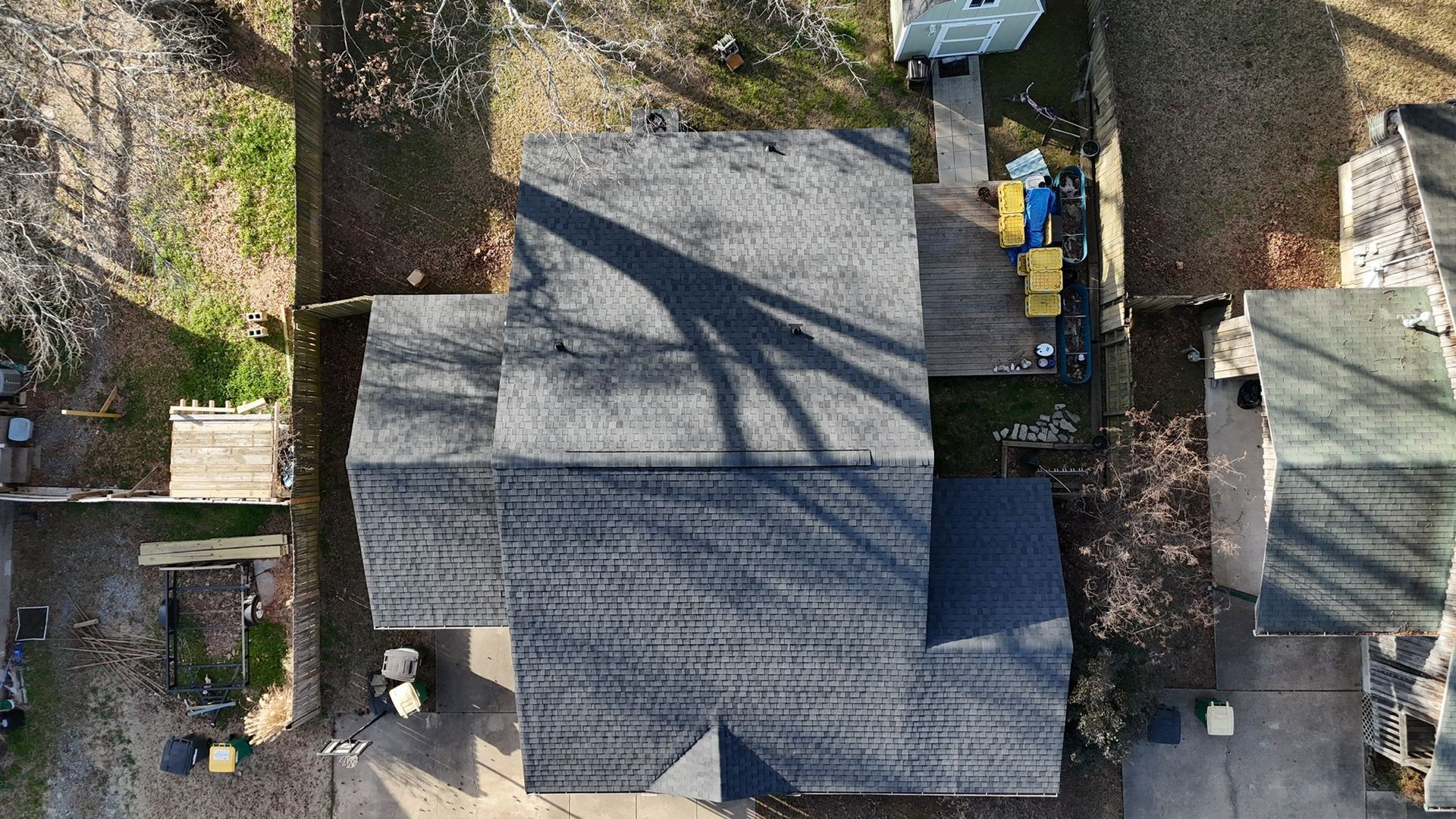 Aerial view of a gray-shingled residential roof with a large tree shadow cast across it, surrounded by trees and a yard.