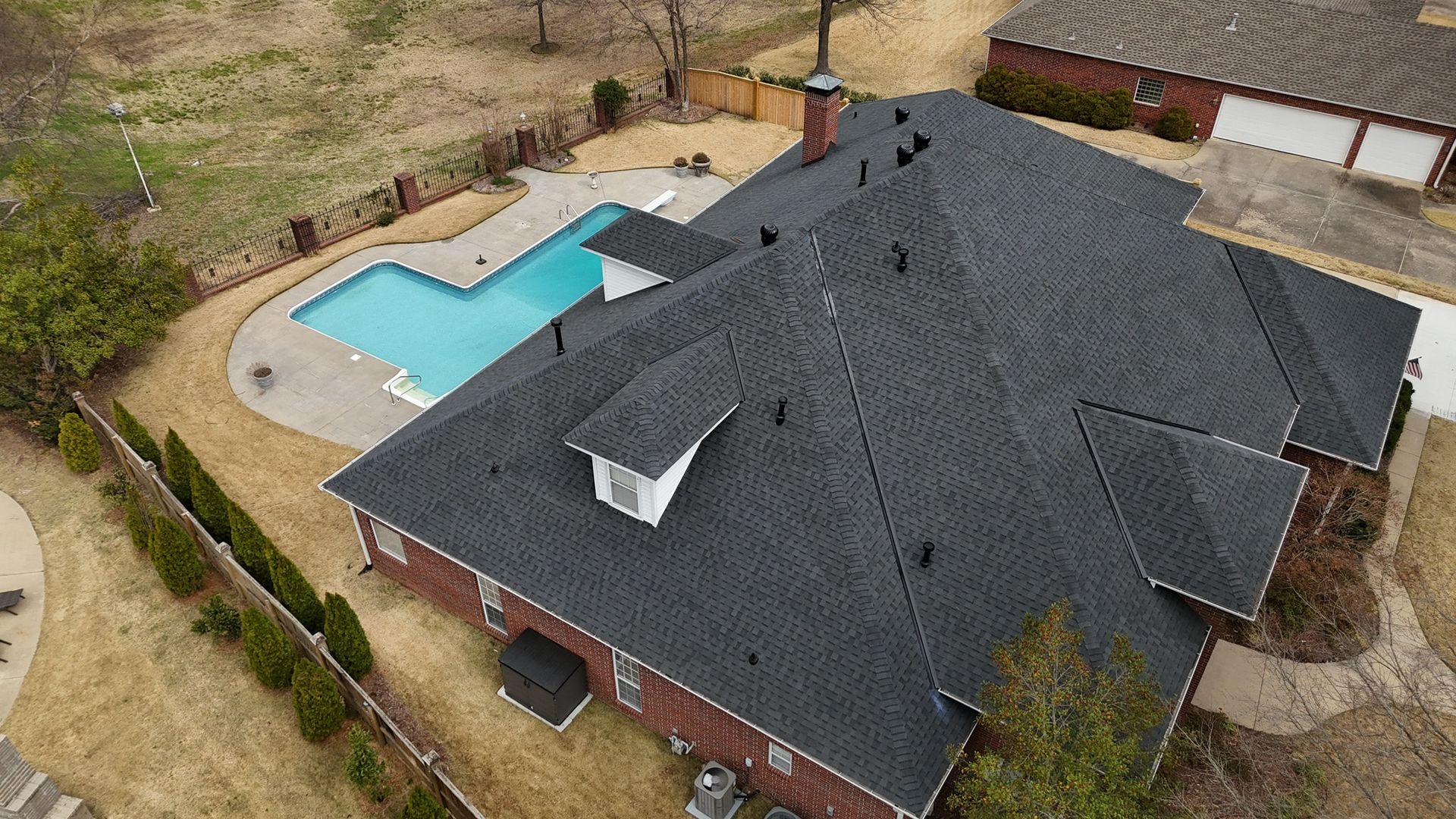 An aerial view of a brick house with a dark shingled roof, a chimney, a dormer, and a backyard swimming pool.