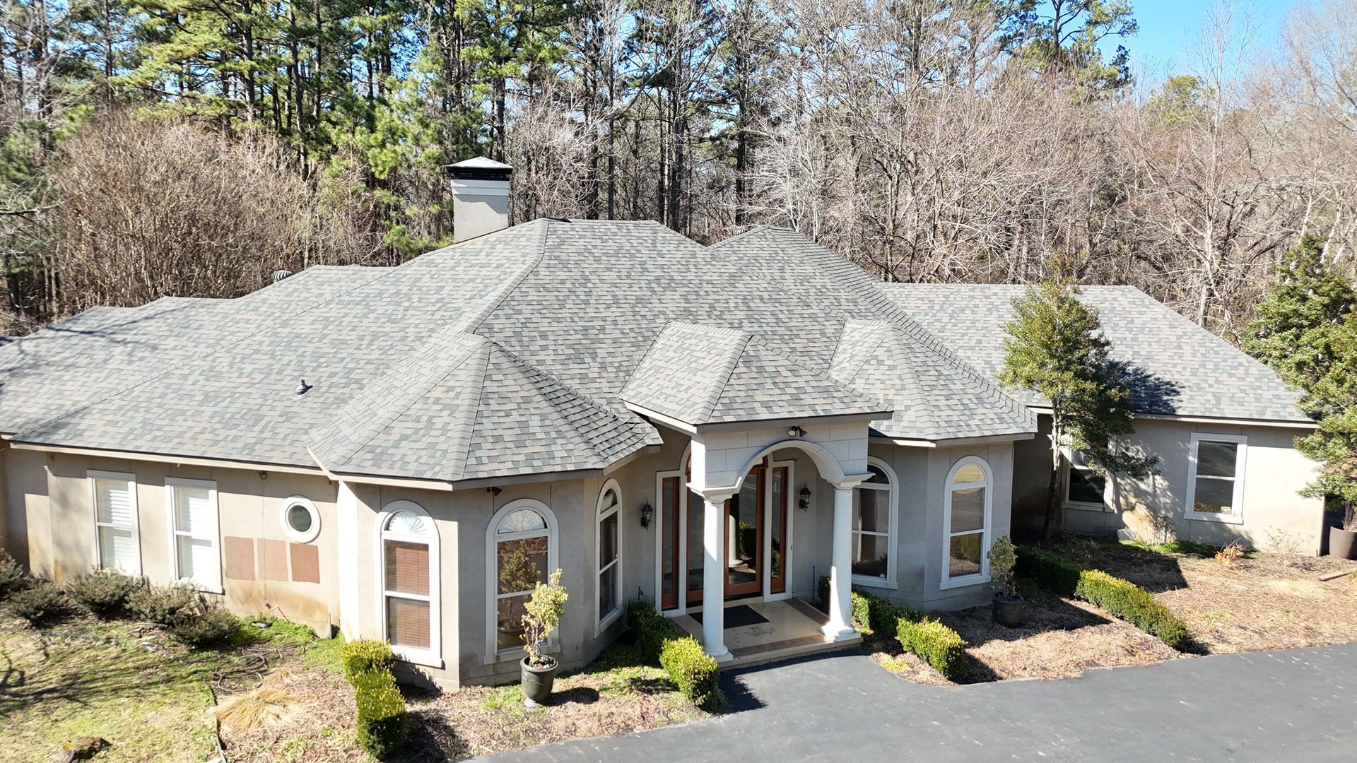 A single-story, beige stucco house with a gray shingled roof, arched front entryway, and a driveway, surrounded by trees.