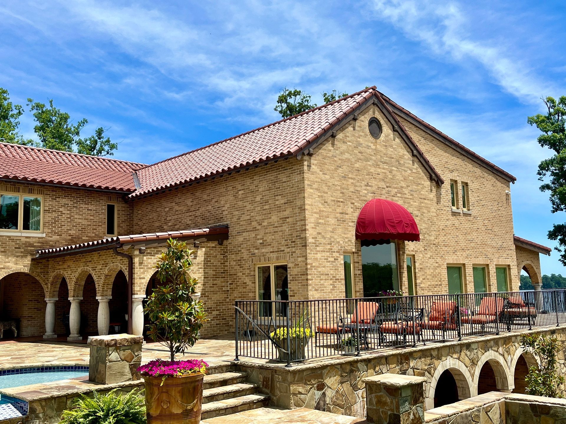 A stone mansion with a red tile roof and patio furniture overlooking a pool on a sunny day.