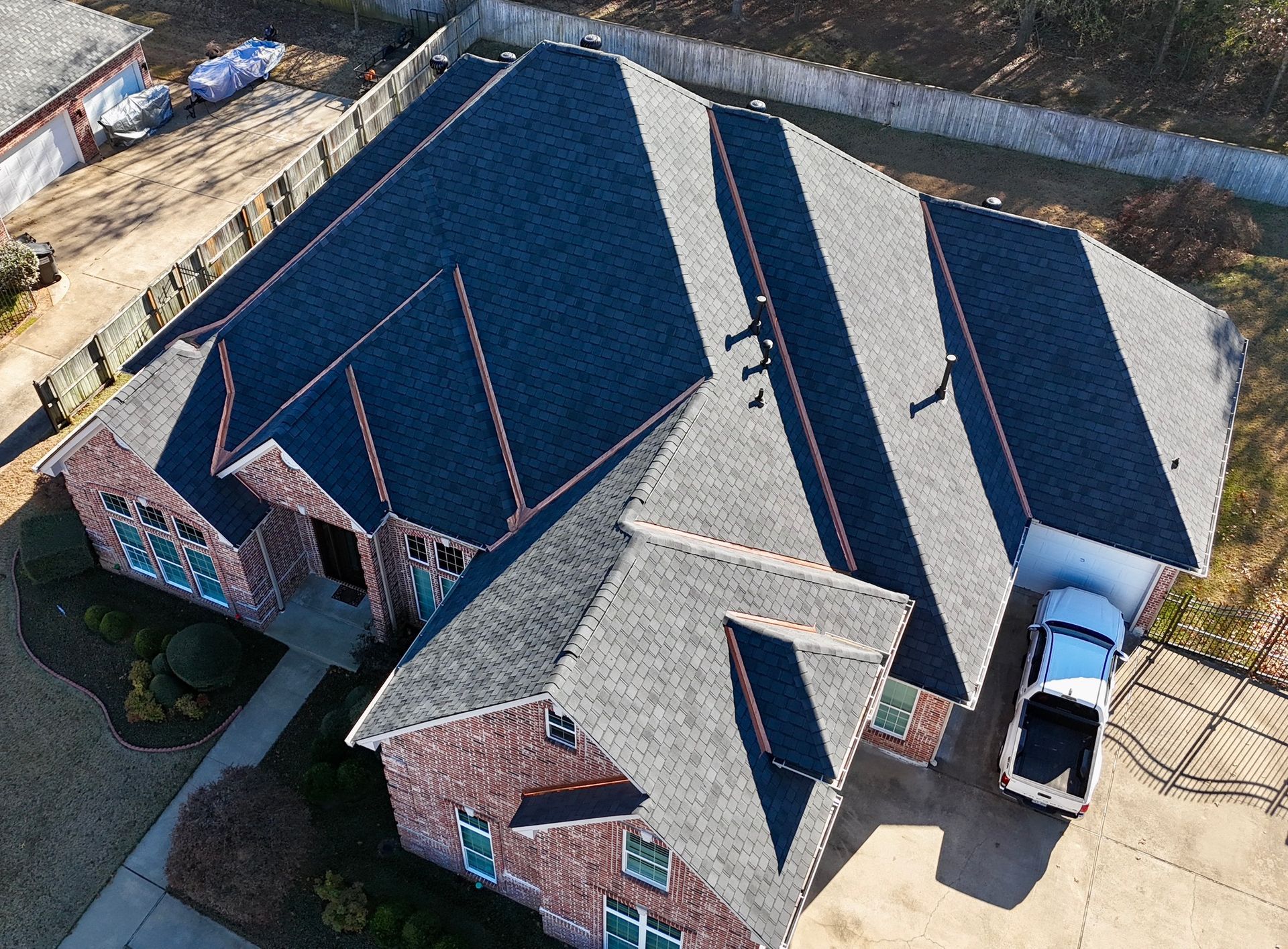 An aerial view of a brick house with a complex, dark gray shingled roof, featuring several peaks and a driveway.