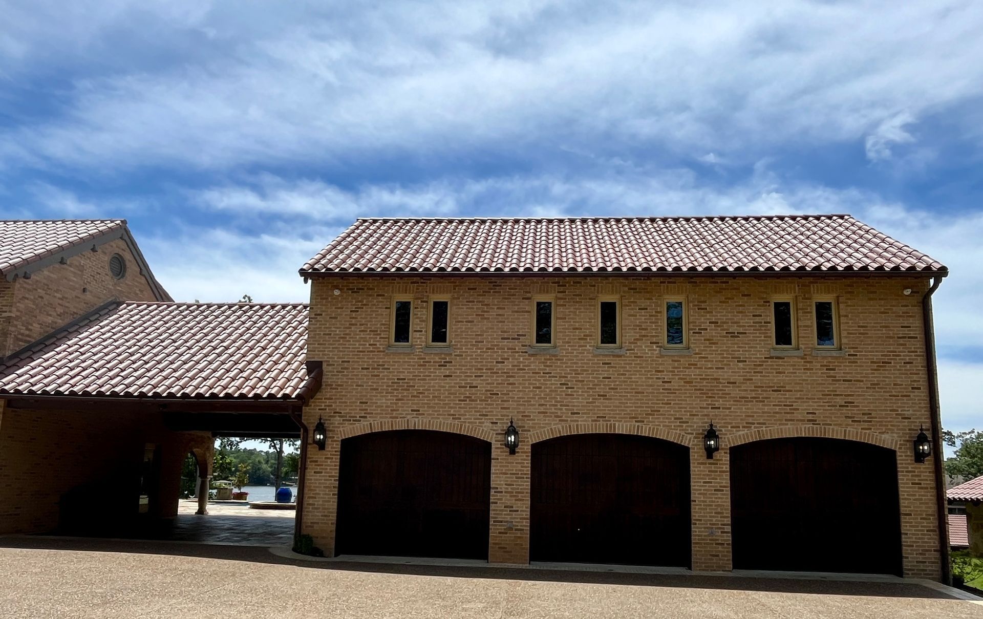 A three-car tan stone garage with a tiled roof and an attached covered walkway under a blue sky with scattered clouds.