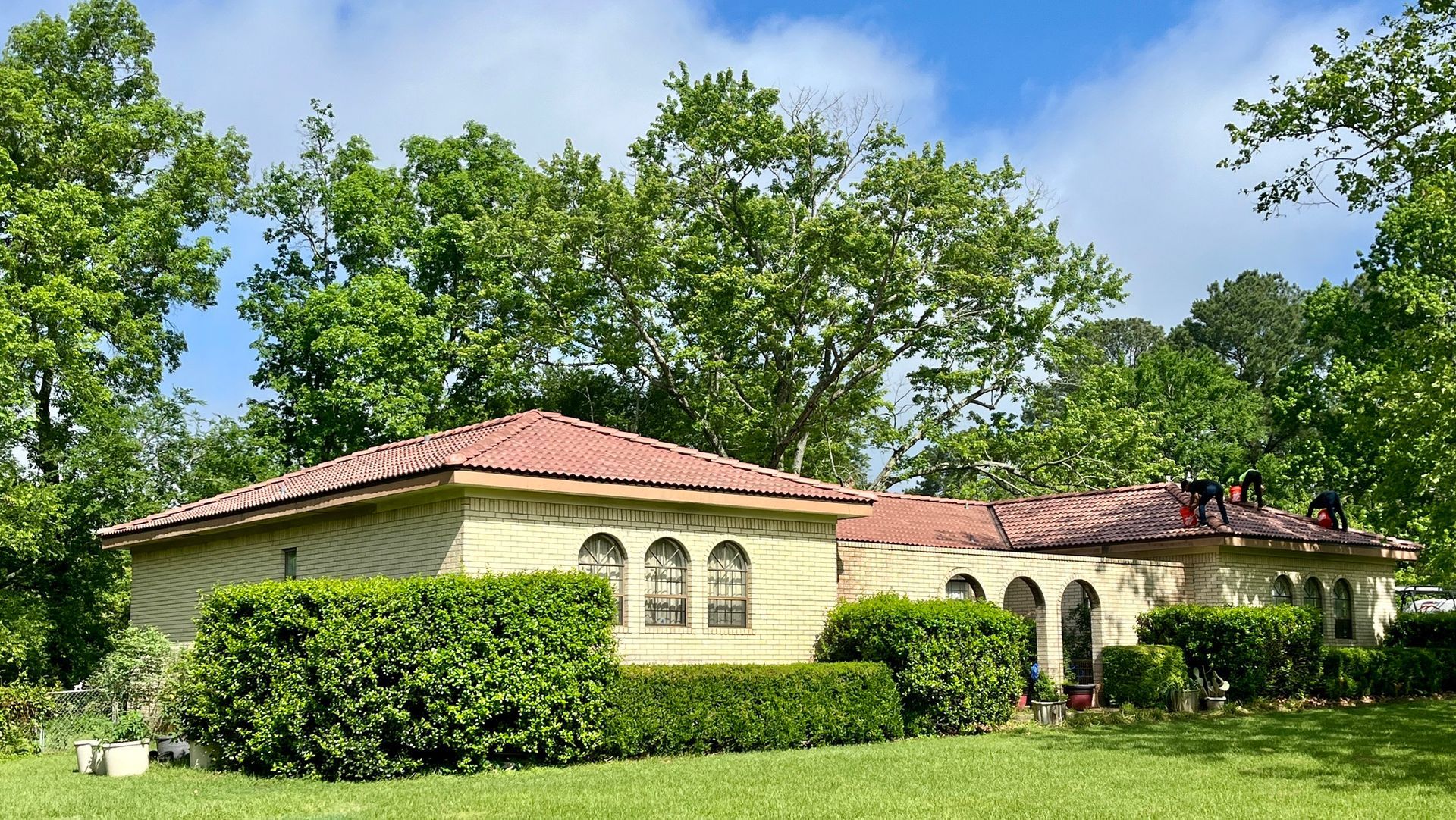 Workers install a new red tile roof on a tan, single-story home surrounded by lush green trees and trimmed hedges.