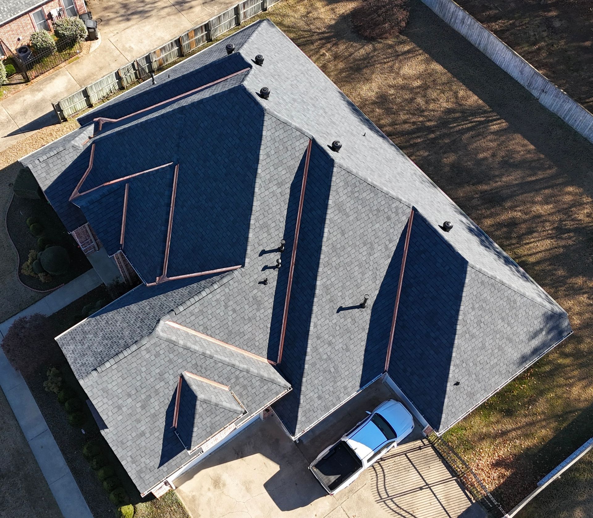 Aerial view of a residential house with a dark gray shingled roof, showing multiple roof planes and a white parked truck.