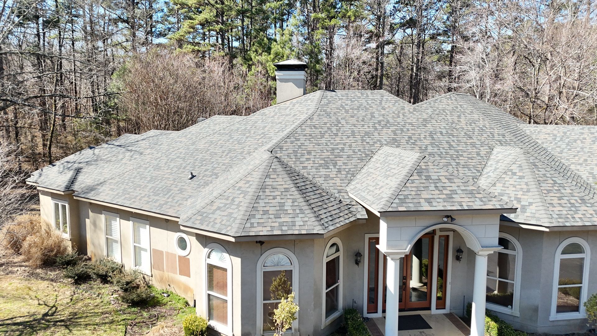 An elevated view of a single-story beige house with a grey shingled roof, surrounded by bare trees in a wooded setting.