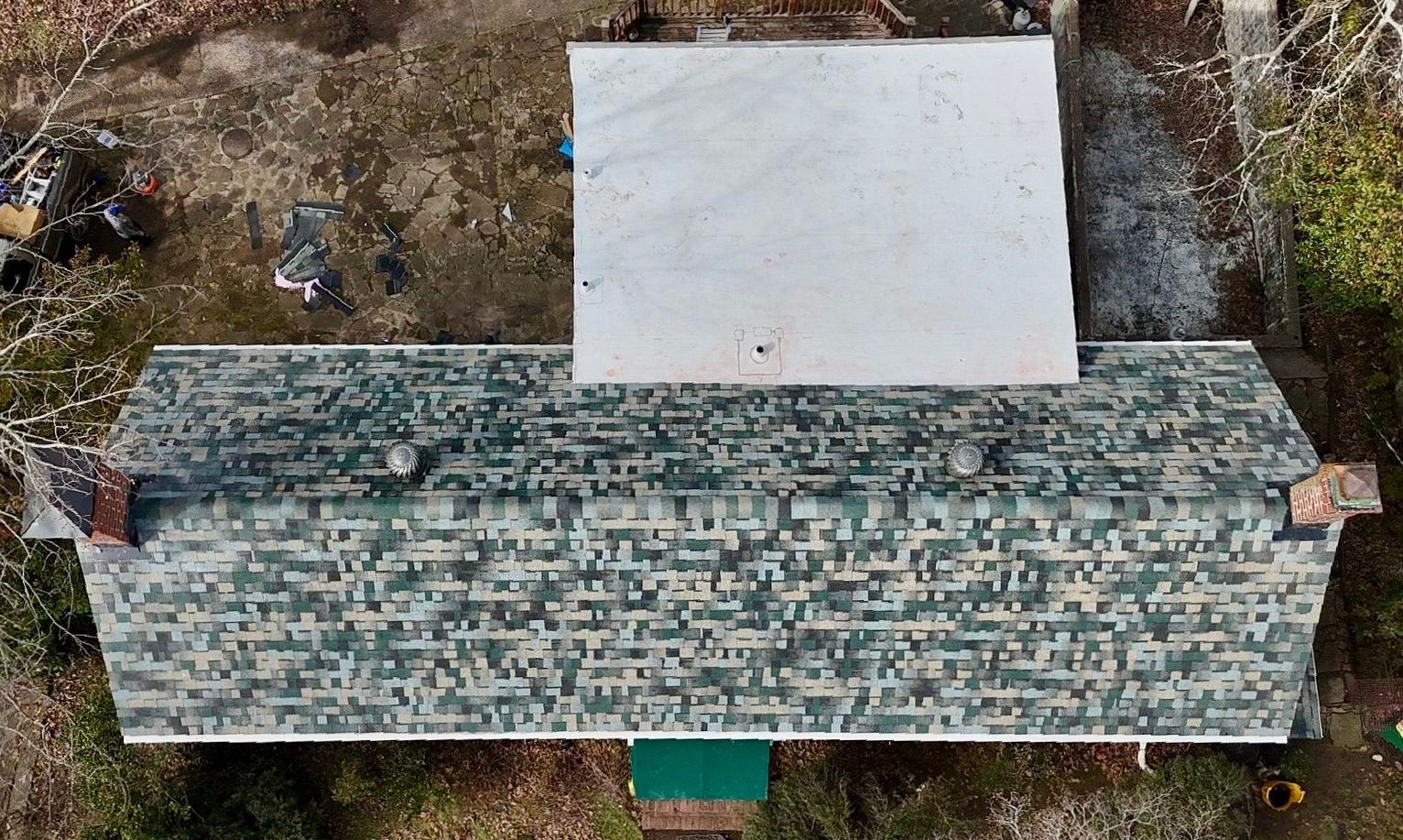 An aerial view of a rectangular house with a blue-green patterned roof and an attached, flat white-roofed section.
