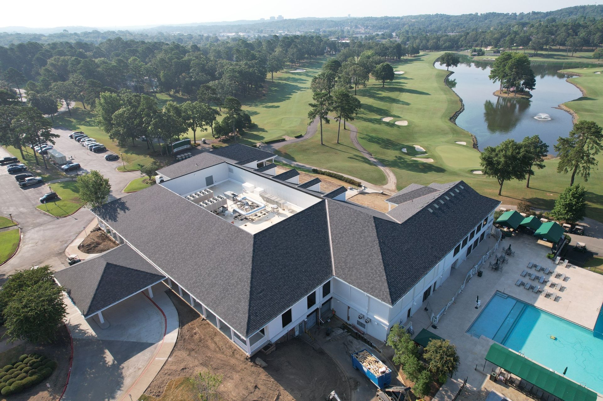 An aerial view of a clubhouse with a dark shingled roof, a swimming pool, and a golf course with a pond in the background.