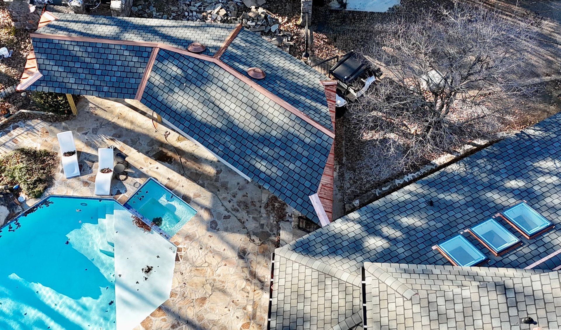 High-angle aerial view of a house with a gray shingled roof and three skylights, next to a blue swimming pool.