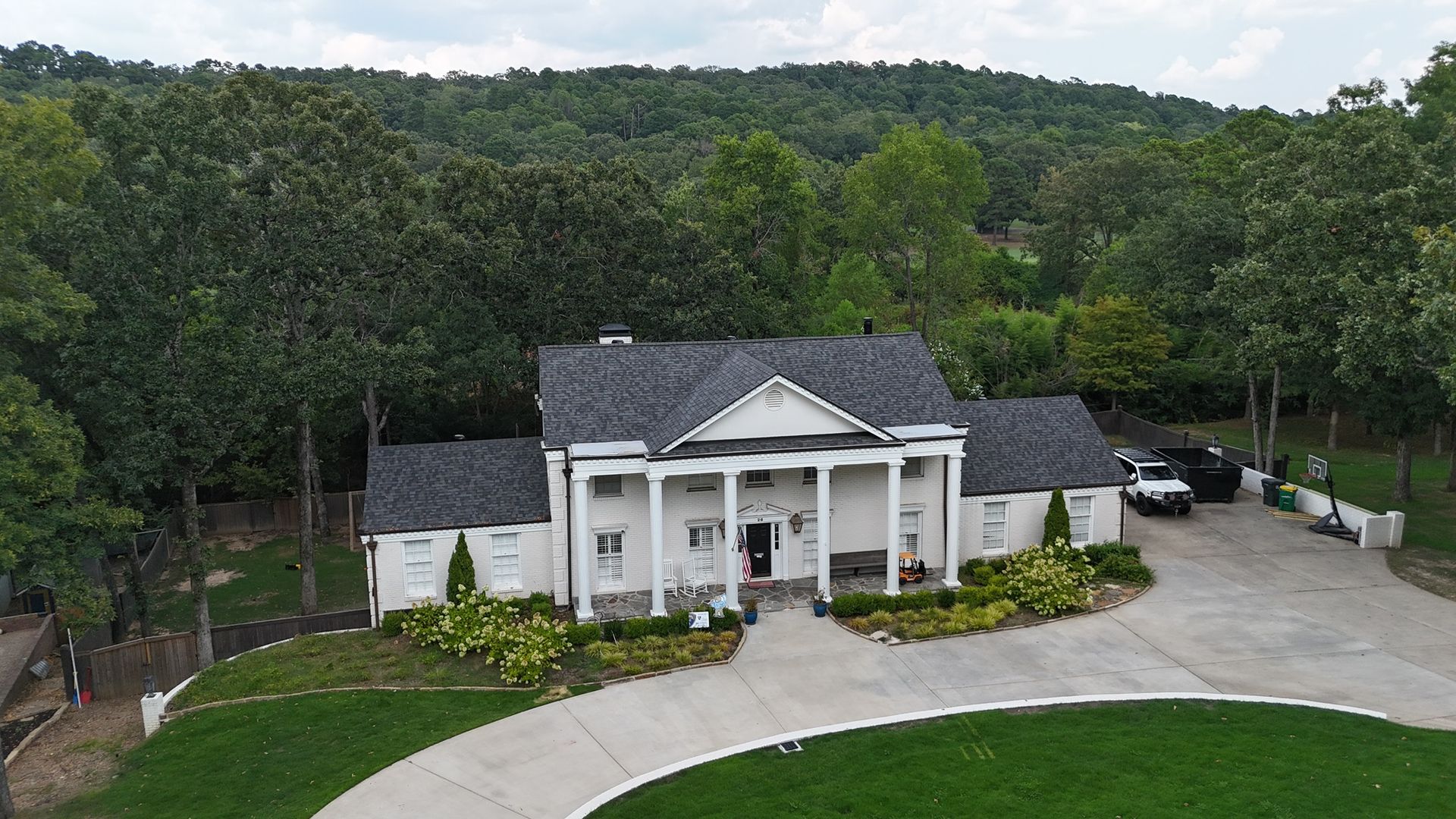 An aerial view of a white, two-story colonial-style house with a gray roof, surrounded by trees and a paved driveway.