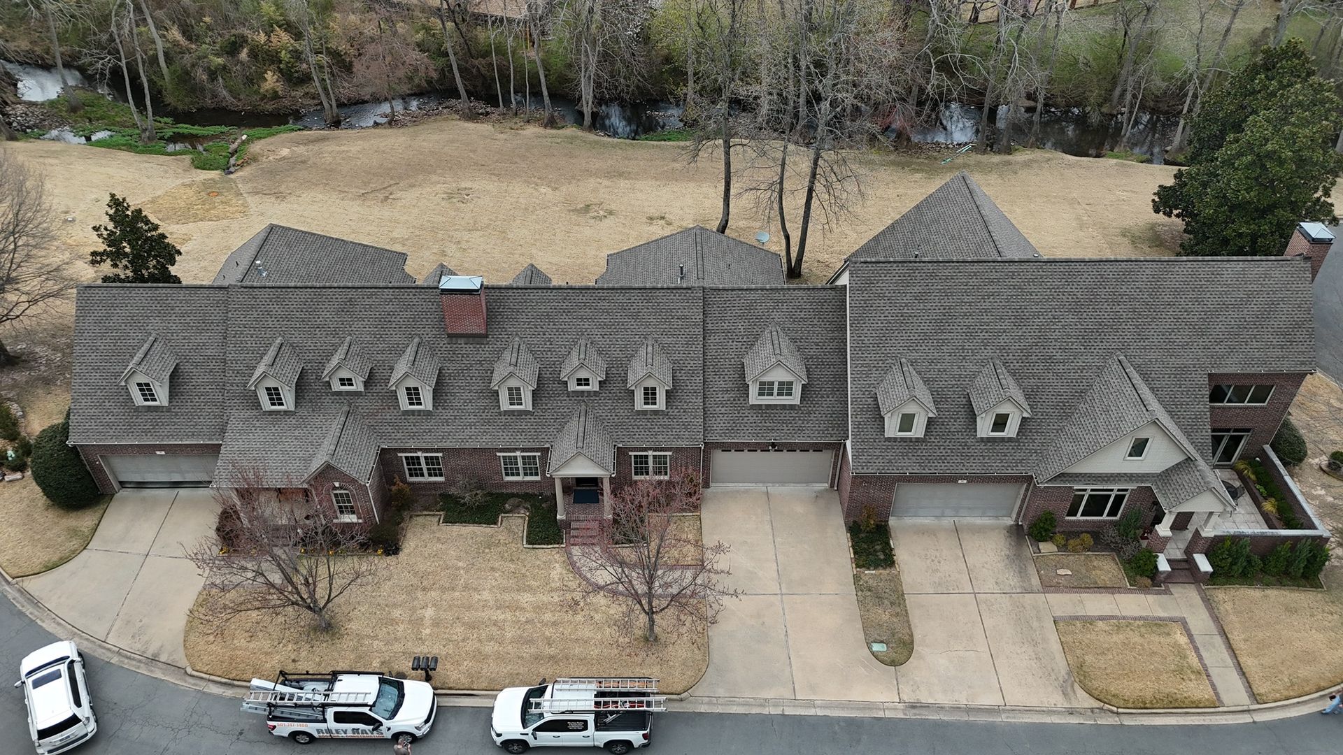 Aerial view of a large brick house with a gray shingled roof, multiple dormer windows, and a driveway in front.