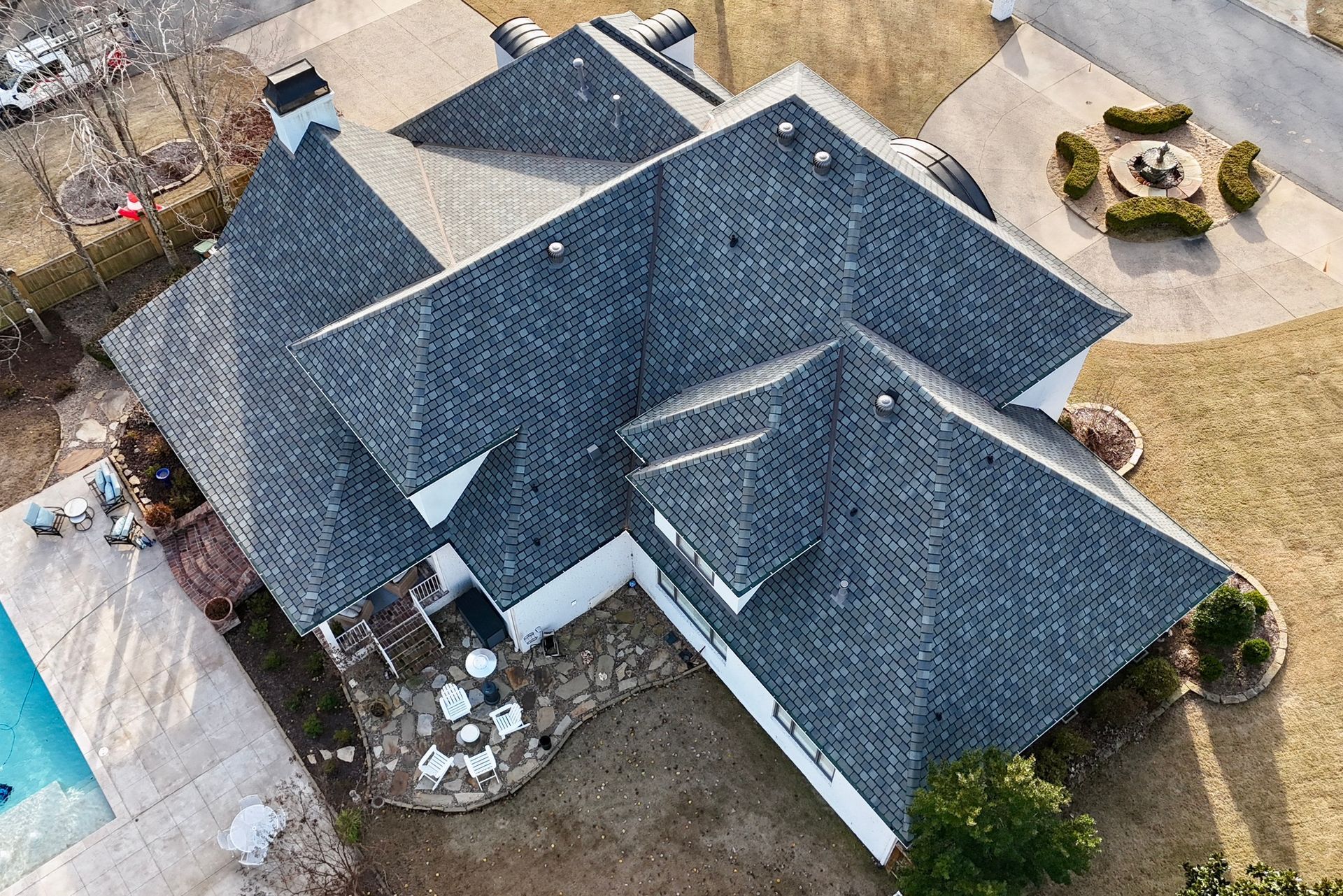 An aerial view of a dark grey shingled residential roof with multiple peaks and a swimming pool in the yard.