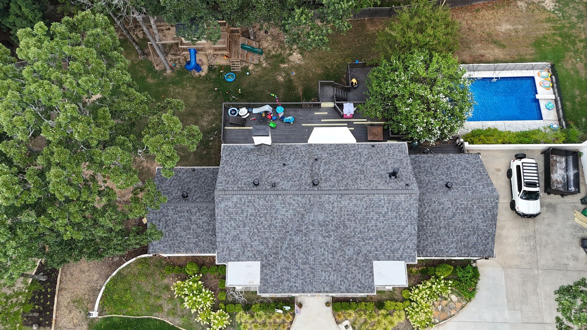 Aerial view of a house with a gray shingled roof, a backyard deck, a rectangular blue pool, and a white vehicle.