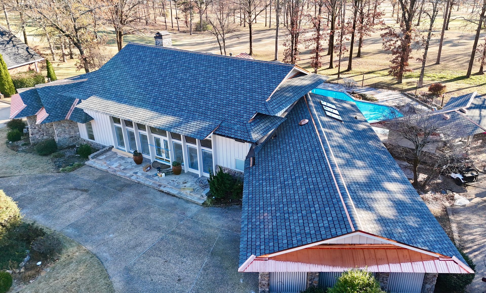 Aerial view of a sprawling house with a blue-shingled roof and copper accents, set in a wooded area with a backyard pool.