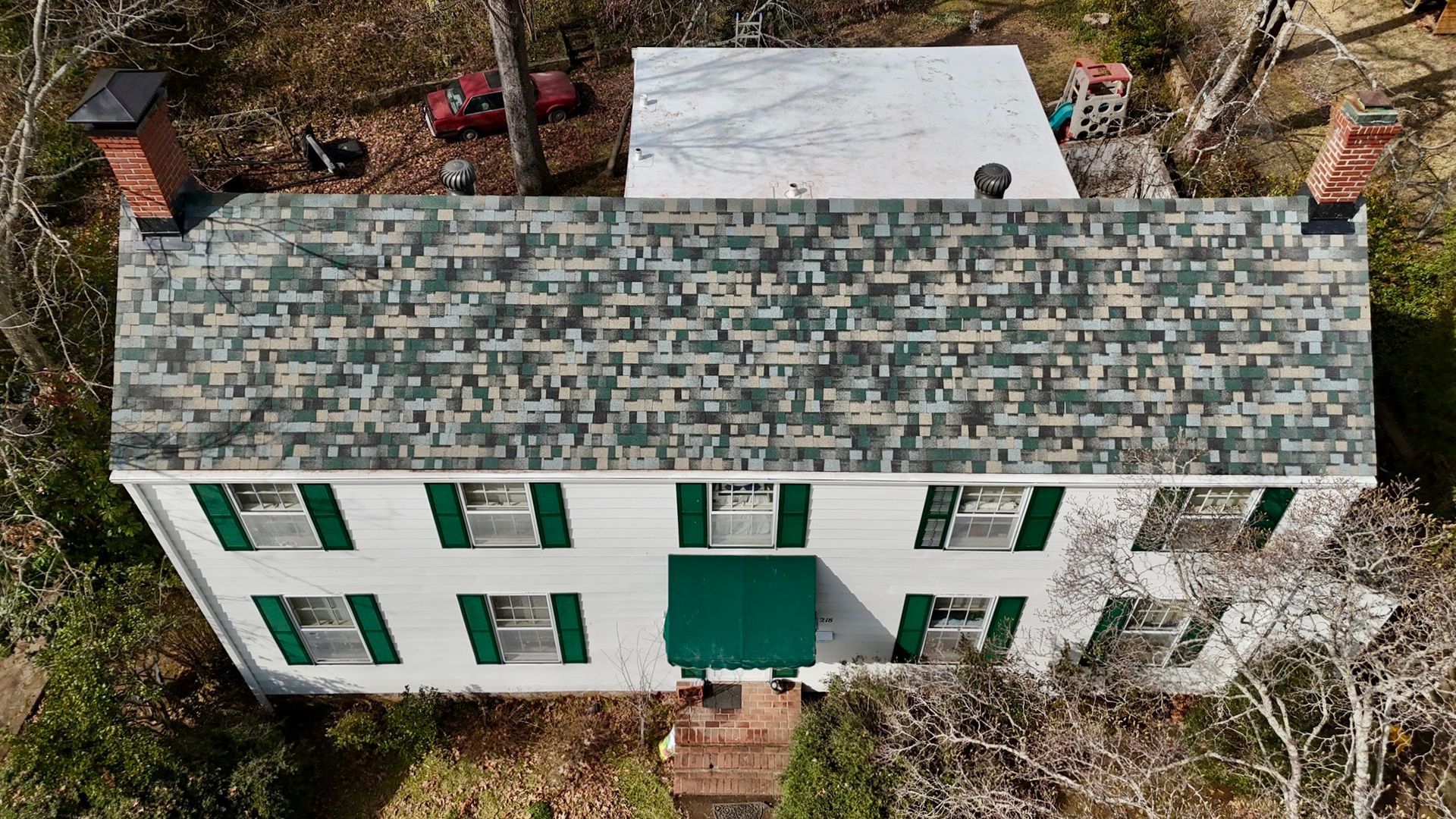 An aerial view of a white, two-story house with green shutters, a patterned shingle roof, and two brick chimneys.