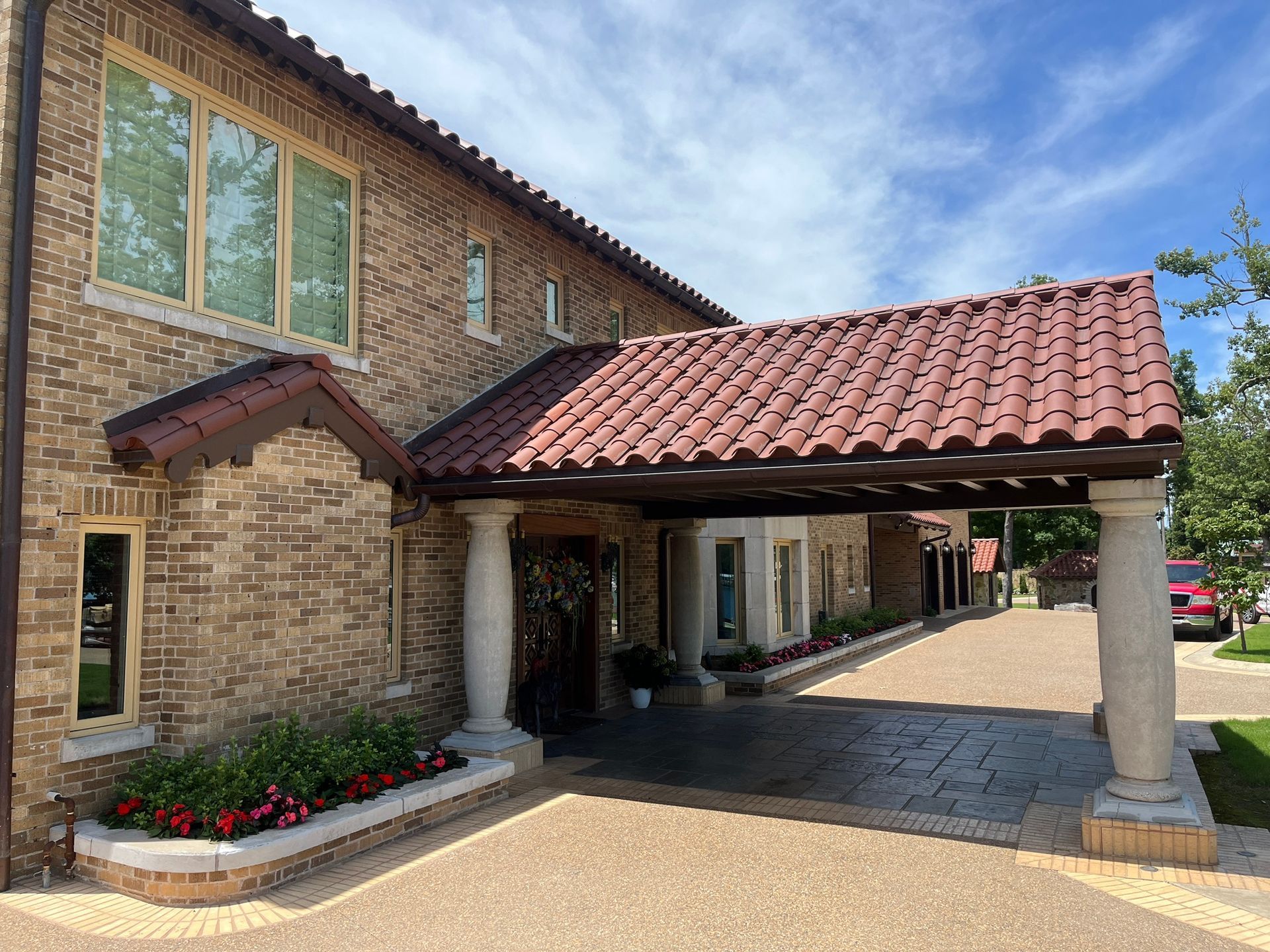 A brick building with a covered portico, stone pillars, a tiled roof, and a flower bed in front on a sunny day.